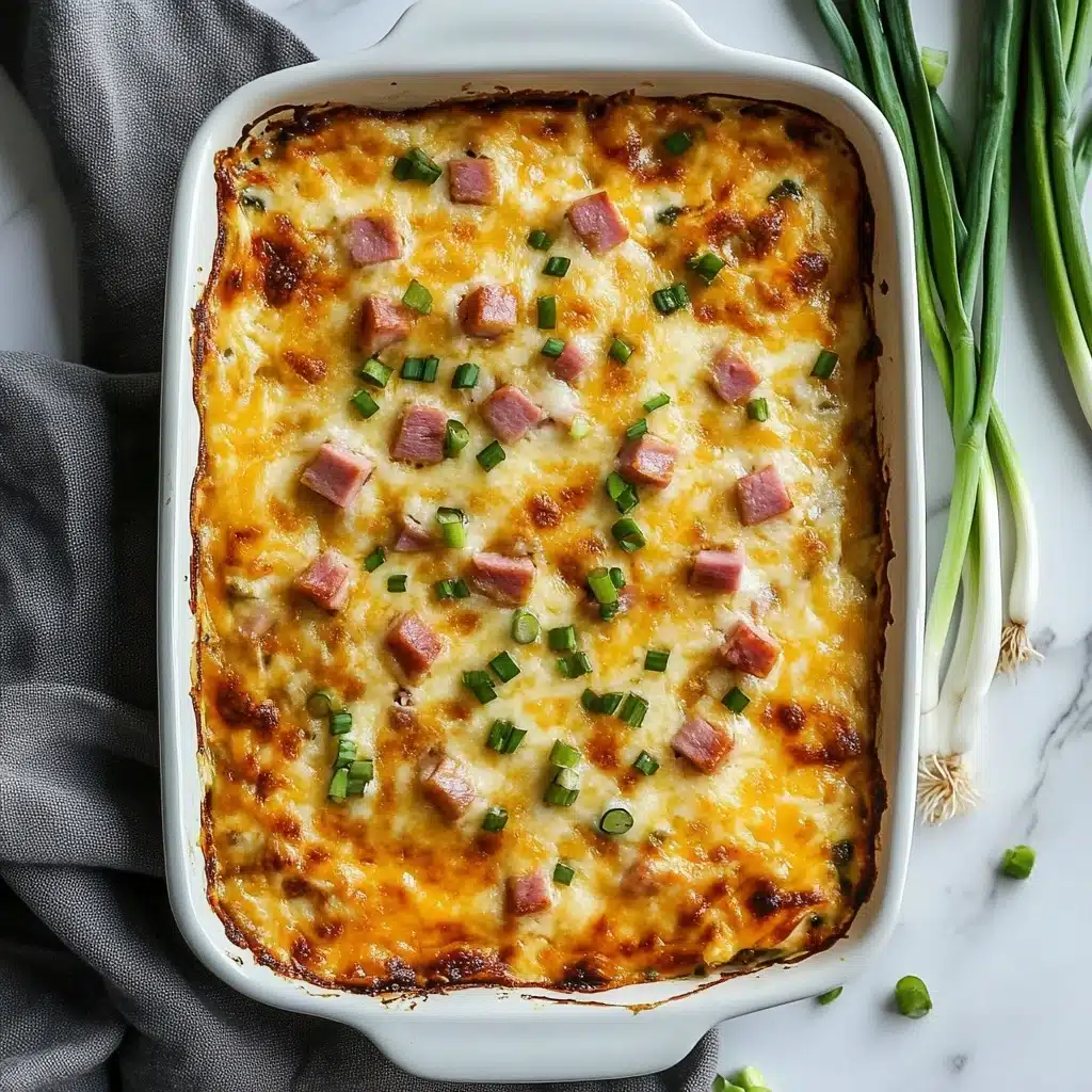 A rectangular white ceramic baking dish filled with a baked casserole showing a creamy top layer with melted and slightly browned cheese, scattered small pink cubes of ham, and green bits of chopped scallions all mixed evenly across the surface; the edges are browned and slightly crispy. The dish is placed on a white marbled surface with a couple of fresh scallions and a gray cloth partially visible beside it. photo taken with an iphone--v 6.1