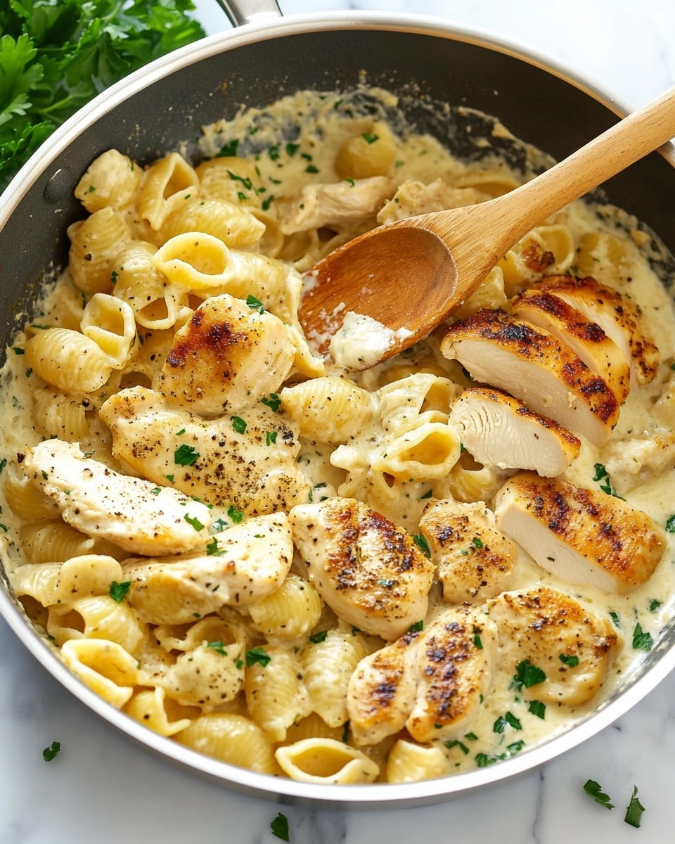 A metal pan filled with creamy pasta shells and sliced, cooked chicken pieces on top. The pasta is covered in a light beige, smooth cream sauce with small green herb bits sprinkled all over. The chicken is golden brown with char marks and pepper seasoning, arranged layered across one side of the pan. A wooden spoon rests inside, leaning across the dish. There is some green parsley decoration near the pan on a white marbled surface. Photo taken with an iphone --ar 4:5 --v 6.1