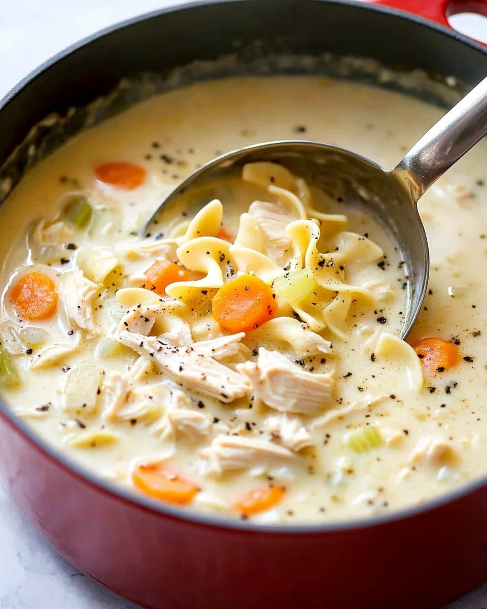 A white bowl filled with creamy chicken noodle soup containing visible layers of shredded light brown chicken, orange carrot slices, pale celery pieces, and short egg noodles all in a thick white broth. On top, a small green sprig of thyme rests for garnish. The bowl sits on a white plate that is layered with a pile of golden round oyster crackers, some scattered around the plate’s edge. A silver spoon is placed inside the bowl, and the whole setup is on a rustic wooden surface. Photo taken with an iphone --ar 4:5 --v 6.1