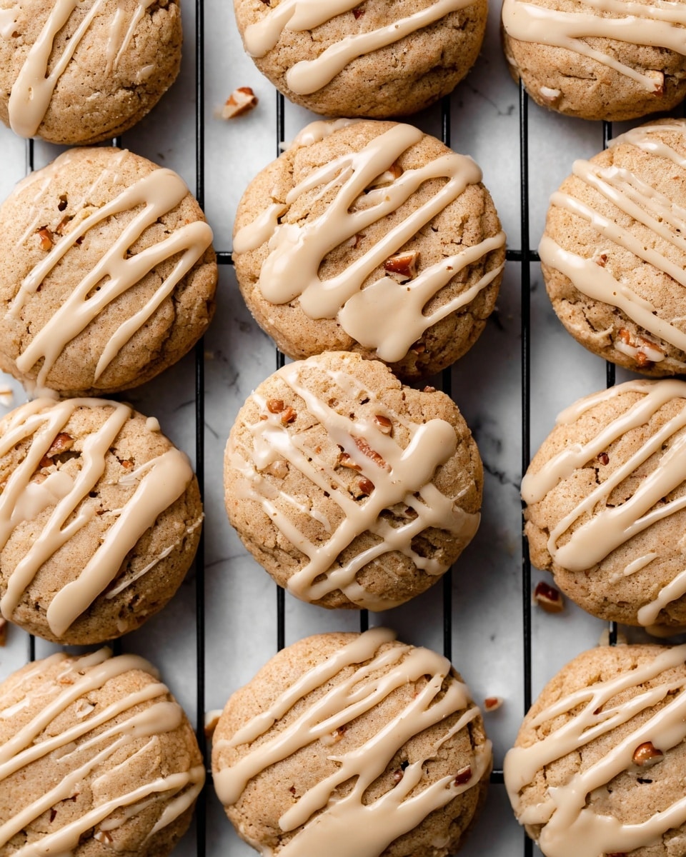 A close-up view of twelve round cookies with a light brown, slightly cracked surface, showing small nut pieces inside. Each cookie has a smooth tan glaze drizzled over the top in uneven lines, some glaze dripping off the edges. The cookies are placed on a black metal rack over a white marbled surface, with minimal shadows. The photo has a bright, clean look. photo taken with an iphone --ar 4:5 --v 7