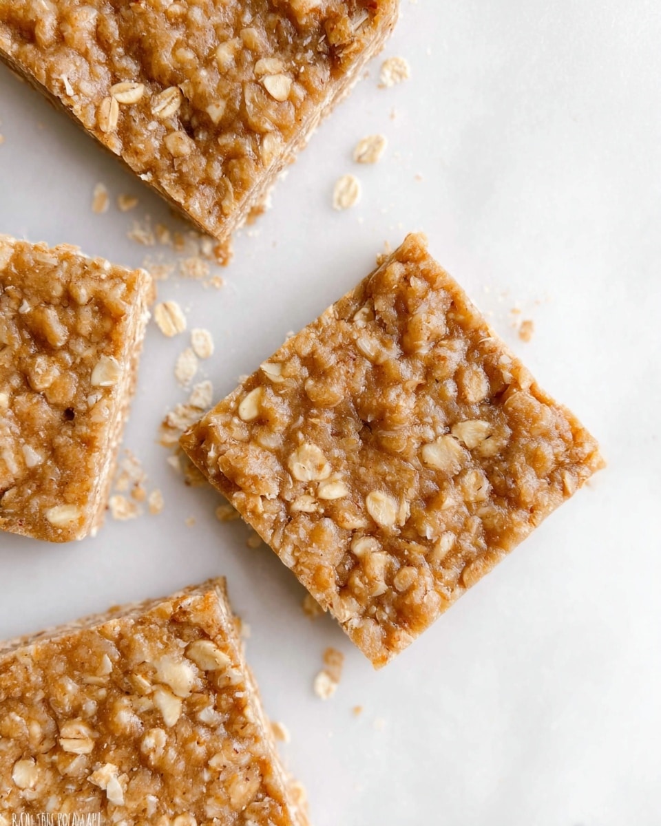 The image shows three square bars stacked on top of each other on a white marbled surface. Each bar is light brown with a rough texture made from oats and other small grains, giving a slightly crumbly look. The bars have a uniform thickness and appear dense, with tiny bits visible inside them. The background is blurred with a soft, light color, creating a clean and simple setting. photo taken with an iphone --ar 4:5 --v 7