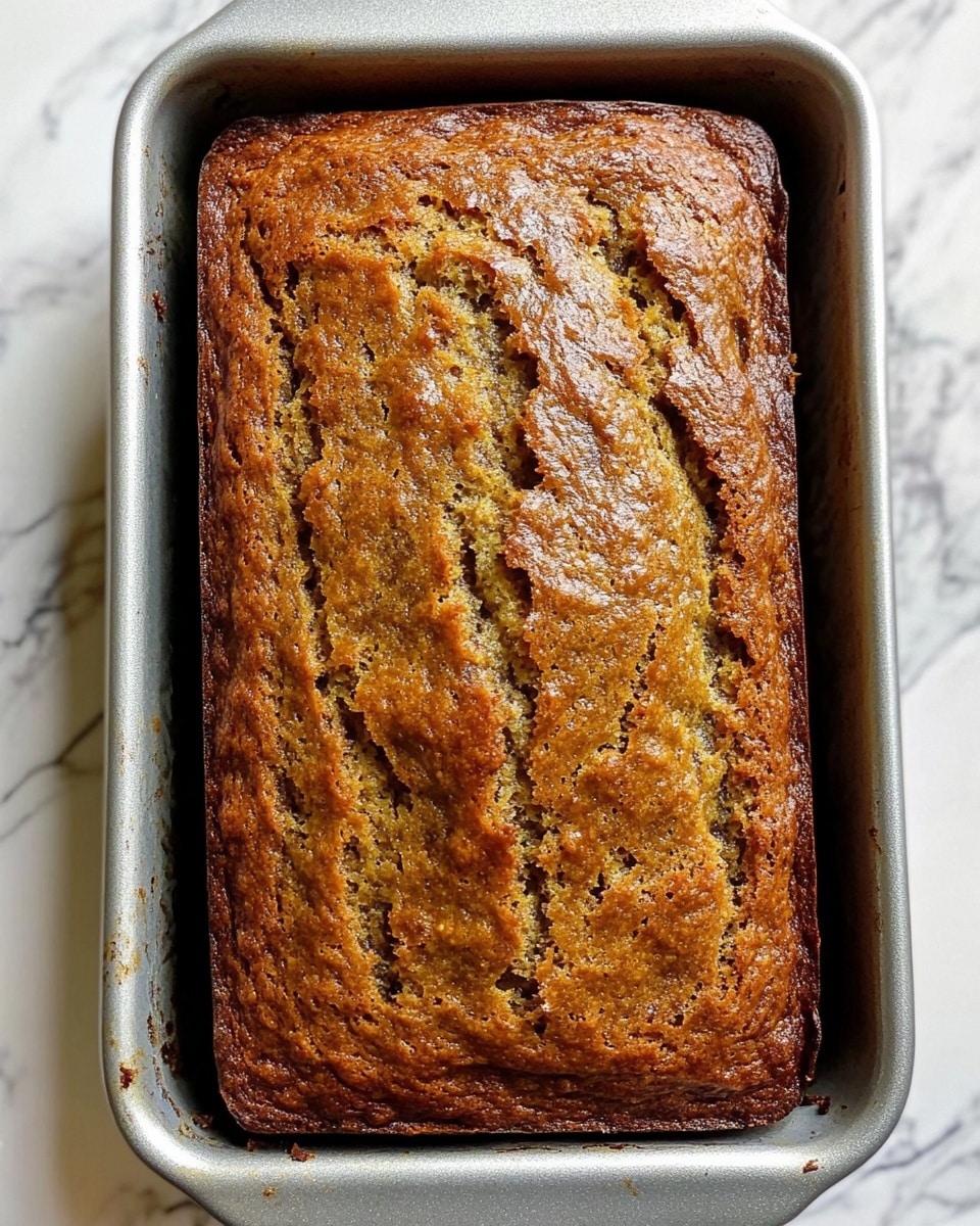 A single, rectangular loaf of banana bread is shown inside a silver metal baking pan. The top crust of the bread is golden brown with a darker browned edge and a cracked, textured surface revealing a softer, lighter banana bread layer underneath. The loaf has an uneven, rustic look with small air bubbles and a slightly rough finish. The pan rests on a white marbled surface. Photo taken with an iphone --ar 4:5 --v 6.1