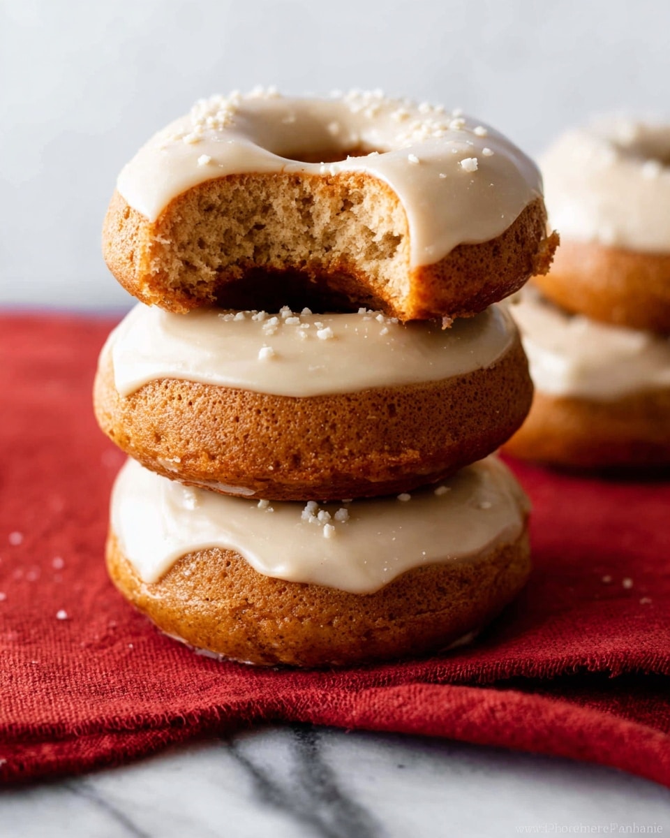 Four donuts are shown on a white ceramic plate with a raised leaf pattern and dark blue glaze. Each donut has one smooth shiny layer of light beige icing on top, and small bits of chopped nuts are sprinkled over the icing. One donut is partly eaten, revealing a soft, golden brown inside layer with a slightly dense texture. Below the plate is a white marbled surface with a piece of a red cloth napkin neatly folded to the right. Photo taken with an iphone --ar 4:5 --v 7