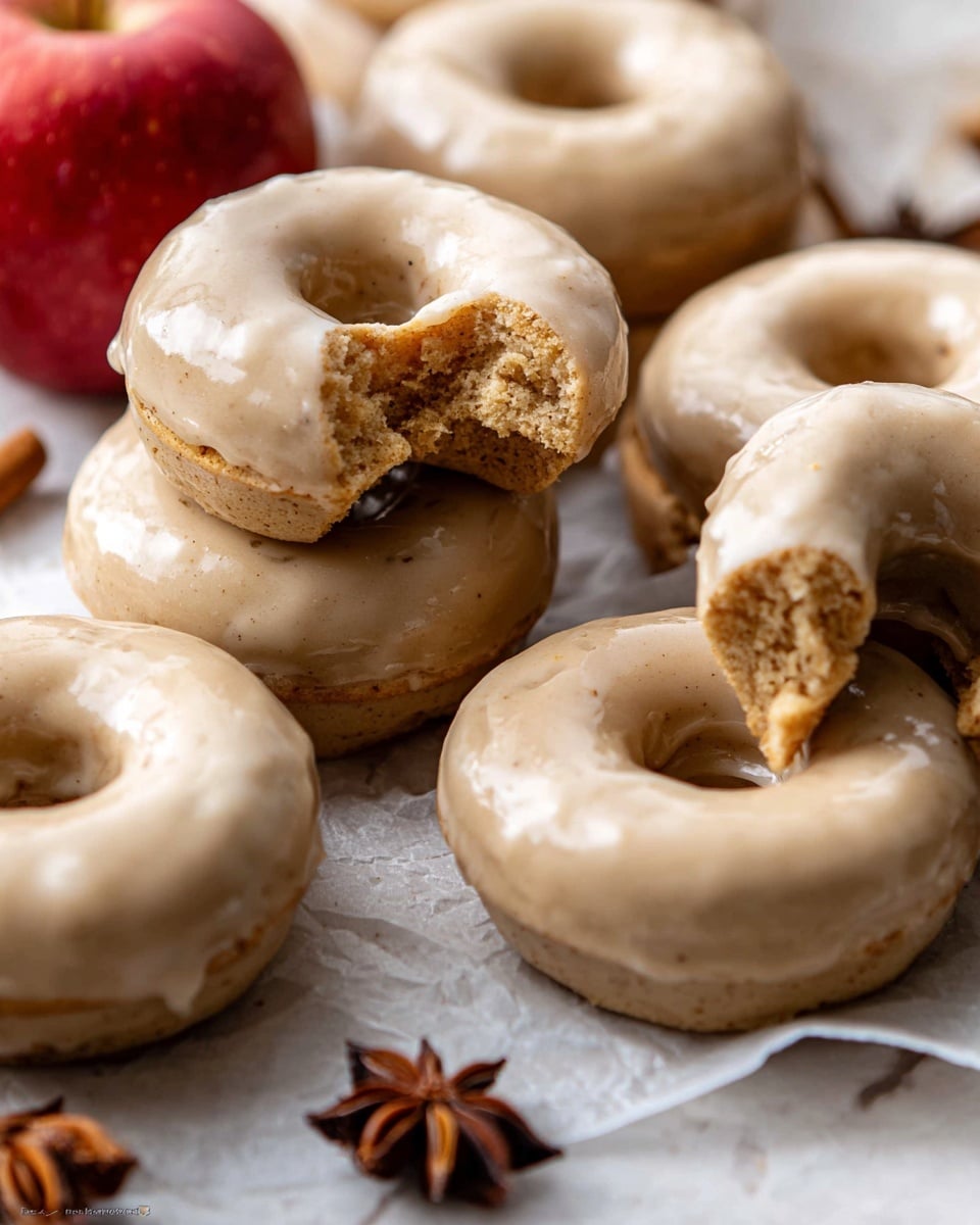 The image shows many round donuts with a light brown glaze that looks smooth and shiny, covering the top and sides. Some donuts are whole, stacked or lying flat, while others are broken or have bites taken out, showing a soft, textured brown inside. An apple with red and yellow skin is placed near the middle right, surrounded by star anise seeds that have a dark brown, spiky shape. A white cup with a red rim is filled with coffee that has a light brown color, and a few star anise seeds float on the surface. All items rest on a white marbled textured surface with a warm, cozy look. photo taken with an iphone --ar 4:5 --v 7