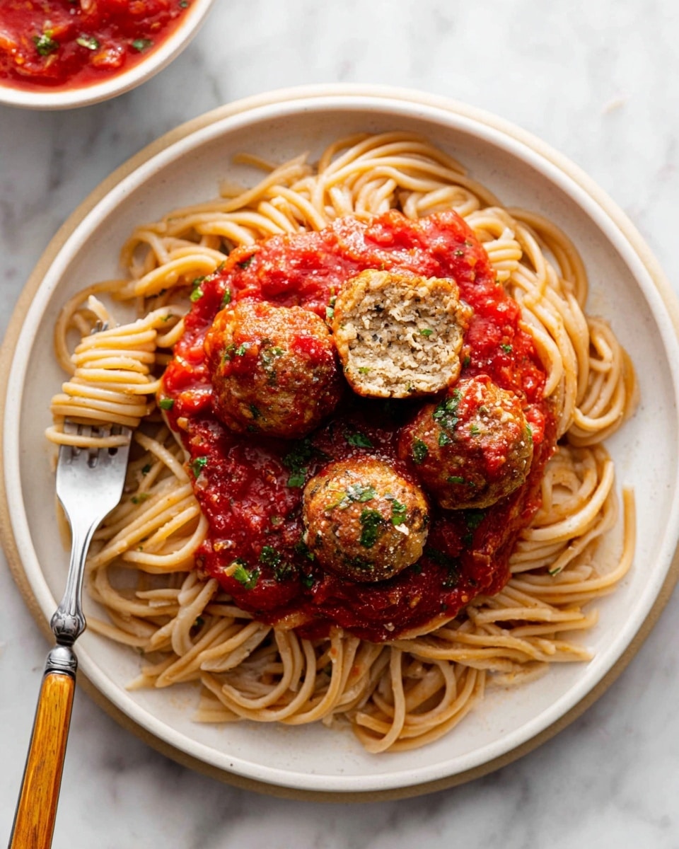 There is a white oval plate filled with about twenty round meatballs evenly coated in a bright red tomato sauce, with small chunks of tomato visible throughout. The meatballs are light brown with a slightly rough texture, and some are sprinkled with small pieces of green parsley. The sauce pools around the bottom of the plate, covering the base and dripping slightly over the edges of some meatballs. A silver spoon is partially submerged in the sauce on the right side of the plate. The plate rests on a white marbled surface, surrounded by small bowls and two glasses filled with a dark red liquid. Photo taken with an iphone --ar 4:5 --v 7
