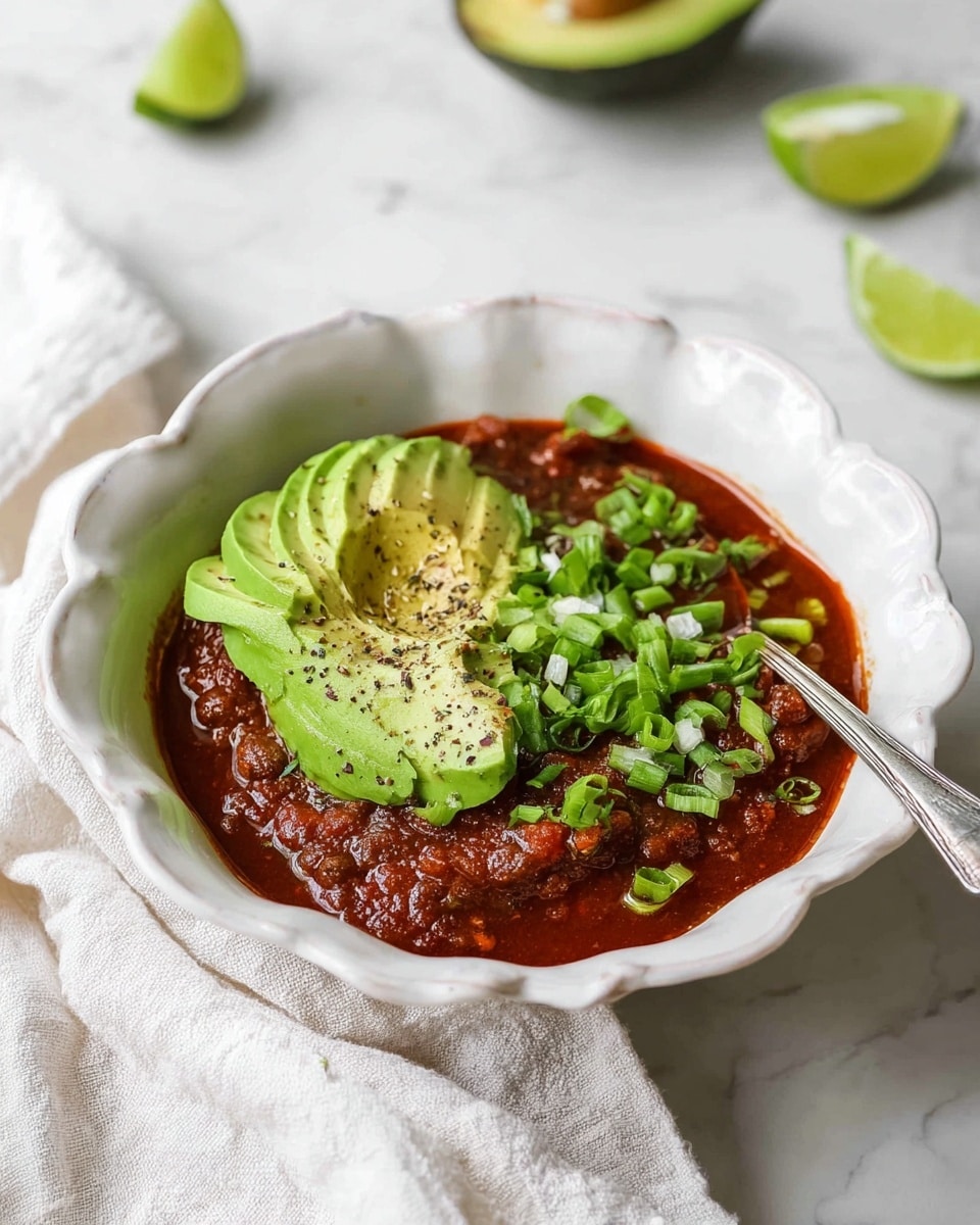 A white bowl with scalloped edges holds a thick, dark red chili with visible beans. On the left side, thin slices of bright green avocado fan out neatly, sprinkled with black pepper. On the right side, vibrant green chopped scallions cover part of the chili. A small, thin lime slice with a green edge is placed near the top center on the chili. The dish sits on a white marbled surface with a halved lime visible at the top left corner. photo taken with an iphone --ar 4:5 --v 7