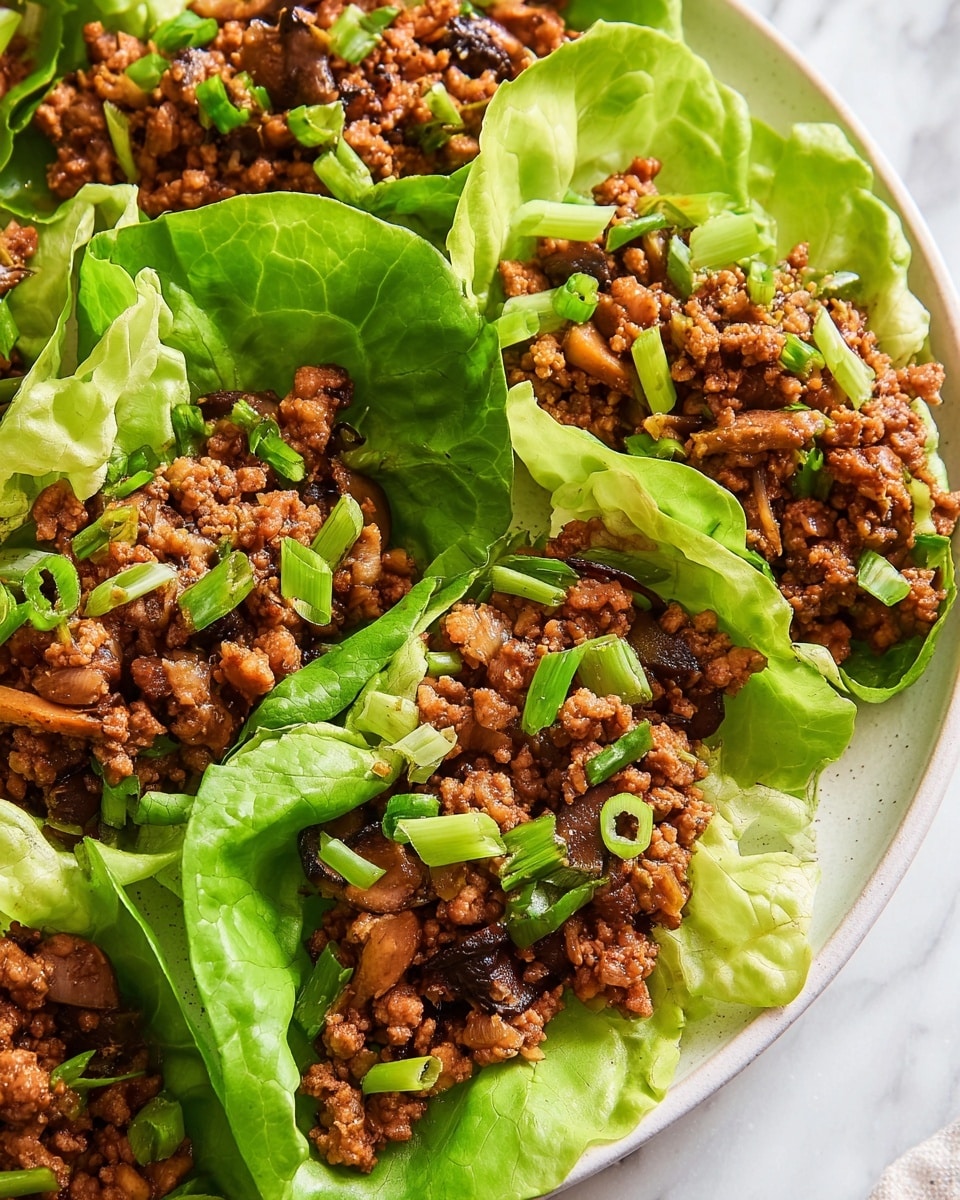 The image shows six lettuce wraps arranged in a circle on a white plate. Each wrap has one bright green lettuce leaf as the base, holding a filling of finely cooked brown minced meat mixed with small pieces of light brown cooked onion and slices of a light yellow ingredient, possibly water chestnuts or a similar vegetable. The filling is topped with small chopped rings of fresh green spring onions, adding a pop of color. The plate sits on a white marbled surface. photo taken with an iphone --ar 4:5 --v 7