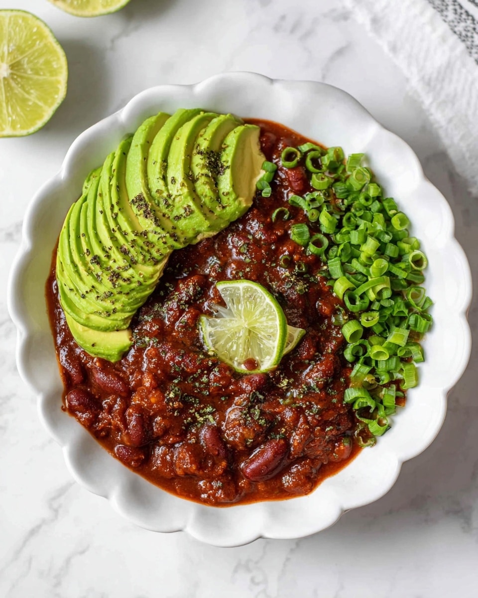 A white bowl with a scalloped edge holds a dark red chili base as the first layer, topped with a large slice of bright green avocado sliced thinly and fanned out on one side. On top of the chili and next to the avocado, there is a thin round slice of lime partially soaked in the chili. The dish is sprinkled with chopped green onions and small bits of white cheese, with a light dusting of black pepper visible on the avocado. A silver spoon rests inside the bowl, leaning on the edge and touching the lime slice. The bowl sits on a white marbled surface with some scattered chopped green onions, lime halves, and cheese crumbs around it. photo taken with an iphone --ar 4:5 --v 7