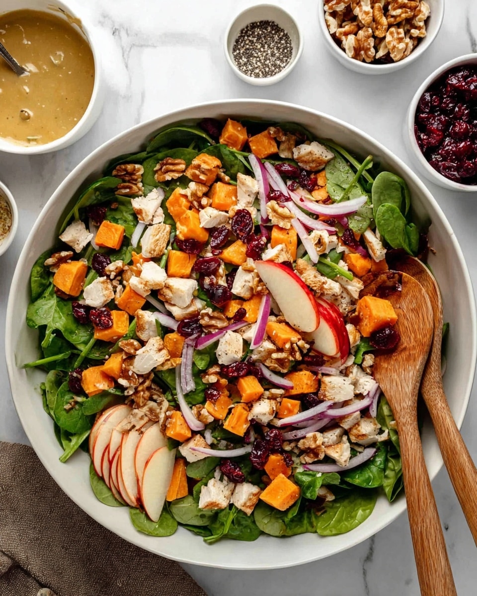 A large white bowl filled with a colorful salad showing many layers; at the bottom there is a bed of bright green fresh spinach leaves, topped with small cubes of orange sweet potato and white apple pieces with red skin visible; scattered on top are pieces of cooked light beige chicken, thin slices of purple-red onion, and dark red dried cranberries; also layered are small chunks of light brown walnuts adding texture. The salad is arranged casually with two wooden spoons resting inside the bowl on the right side. Surrounding the bowl are small white dishes holding walnuts and pepper, a glass jar with a light beige sauce, a bottle of avocado oil, a green-capped bottle of white dressing, and a small bowl of dried cranberries, all set on a white marbled surface. Photo taken with an iphone --ar 4:5 --v 7