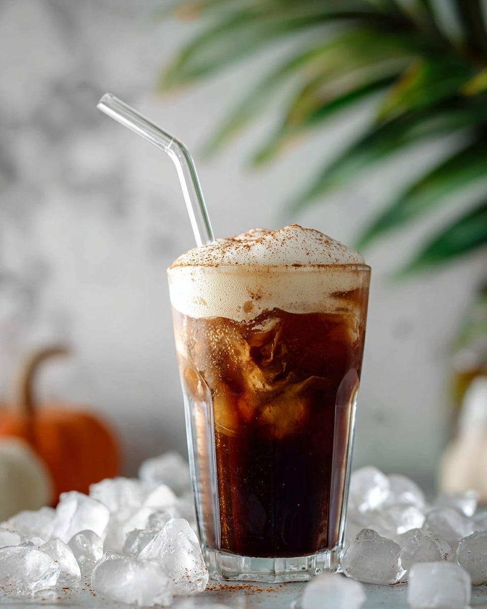 A clear glass filled with dark brown iced coffee layered with light brown ice cubes and topped with a thick, creamy off-white foam sprinkled with fine brown powder, a clear bent straw sticking out from the foam. The glass is set on a bed of scattered ice cubes against a background of blurred green leaves and a white marbled surface. Photo taken with an iphone --ar 4:5 --v 7