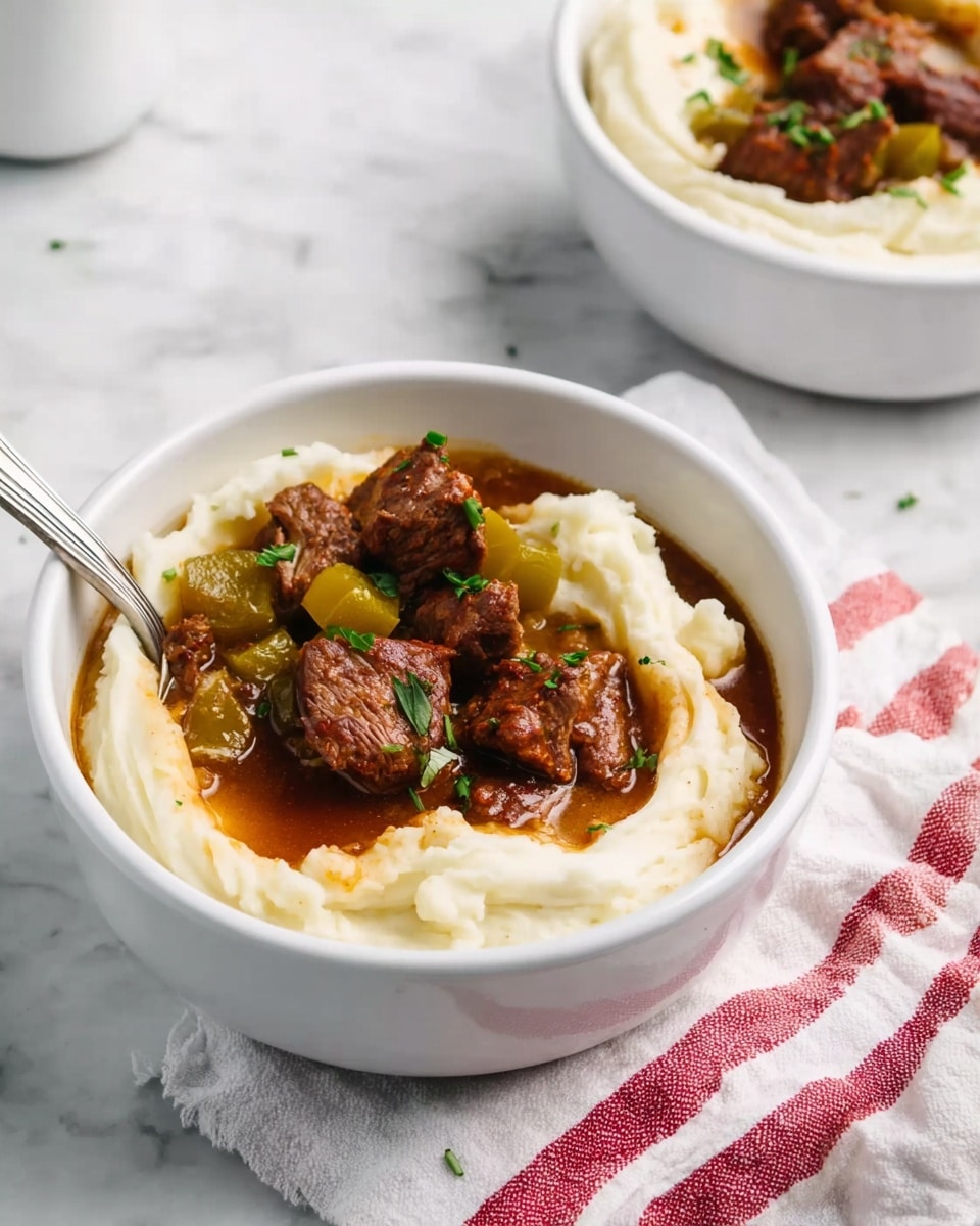 The image shows a white bowl filled with a base layer of creamy mashed potatoes with a smooth texture and slight swirls. On top, there are several pieces of tender brown meat and yellow-green peppers scattered unevenly. A rich brown gravy is drizzled over the meat and mashed potatoes, adding a shiny finish. A silver spoon is partially placed inside the bowl, resting on the mashed potatoes. The bowl sits on a white marbled surface, next to a folded cloth with red stripes. In the background, there is another white bowl with the same dish slightly out of focus. photo taken with an iphone --ar 4:5 --v 7