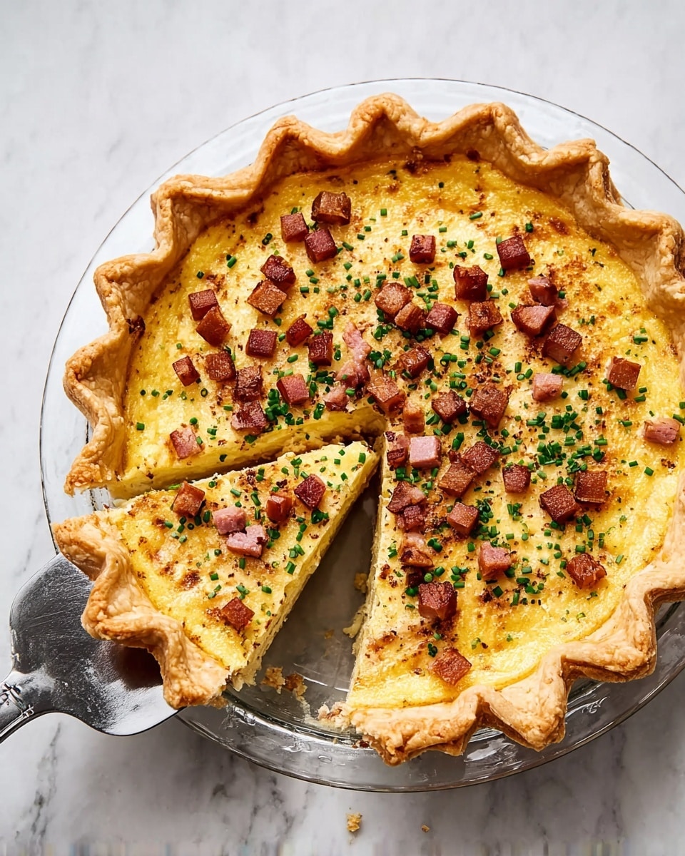A golden baked quiche with a thick, crimped crust sits on a clear glass plate over a white marbled surface. The quiche has one slice removed, showing its smooth, creamy, yellow filling inside. The top layer is sprinkled with small, browned cubes of crispy ham and finely chopped green chives, adding pops of color and texture. A large metal pie server rests beside the plate on the white marbled surface. Photo taken with an iphone --ar 4:5 --v 7