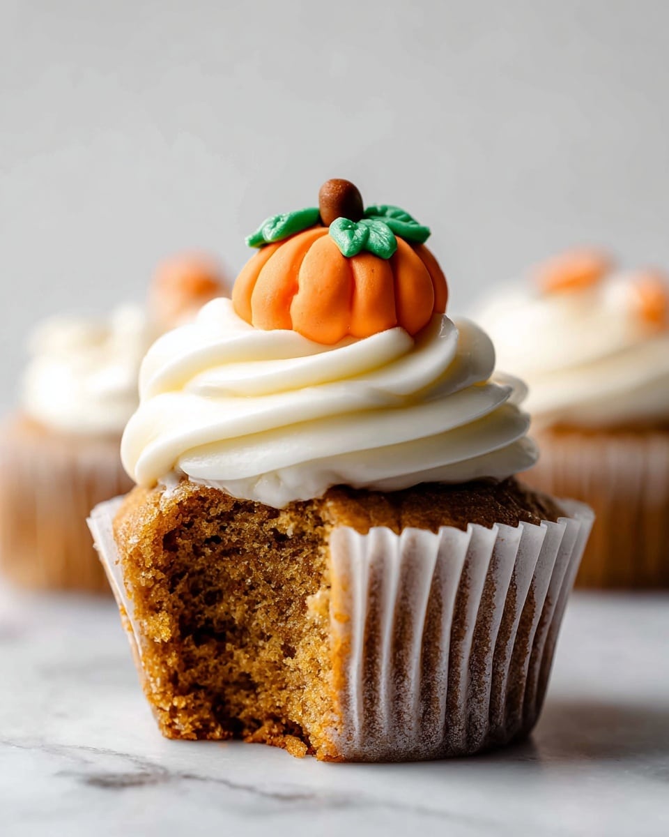 Several brown cupcakes with light beige swirled frosting on top are arranged on a white marbled surface. Each cupcake is wrapped in a paper liner and decorated with a small orange pumpkin-shaped candy with green leaves placed at the center of the frosting. One cupcake lies in the background, blurred, while the closest cupcake is in sharp focus. A small orange pumpkin candy also lies on the surface in front of the cupcakes. The overall scene has bright, natural lighting. Photo taken with an iphone --ar 4:5 --v 7