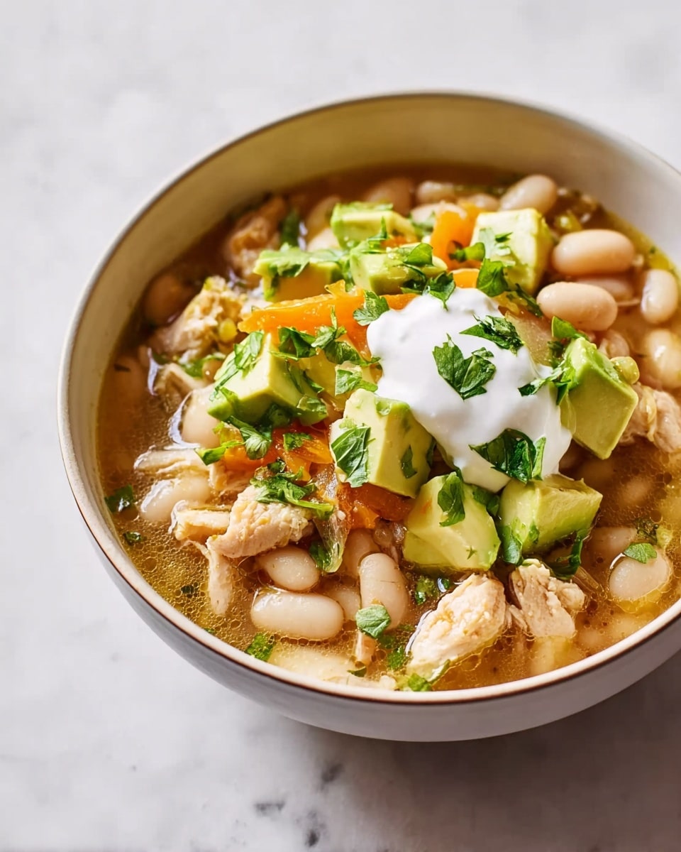 A bowl filled with white beans in a light brown soup, with pieces of cooked meat mixed in, topped with small green avocado cubes, a dollop of white sour cream in the center, and sprinkled with thin yellow cheese strips and chopped green herbs. The bowl is white with a brown rim and sits on a white marbled surface. Photo taken with an iphone --ar 4:5 --v 7