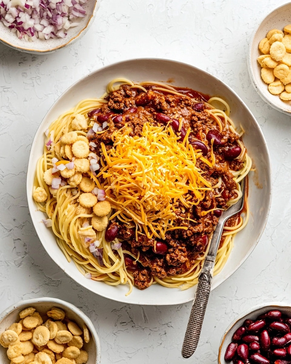 A white plate on a white marbled surface holds a dish with three main layers: the bottom layer is plain spaghetti noodles arranged in a loose pile, the middle layer is chunky chili sauce with red kidney beans spread unevenly over the noodles, and the top layer is shredded orange cheddar cheese sprinkled over everything, along with small round oyster crackers scattered on top and mixed in. To the left of the plate are two small bowls, one with diced white onions and the other with oyster crackers, and to the right are two bowls filled with red kidney beans and shredded cheddar cheese. A fork rests diagonally on the plate, partially on the noodles and chili. Photo taken with an iphone --ar 4:5 --v 7