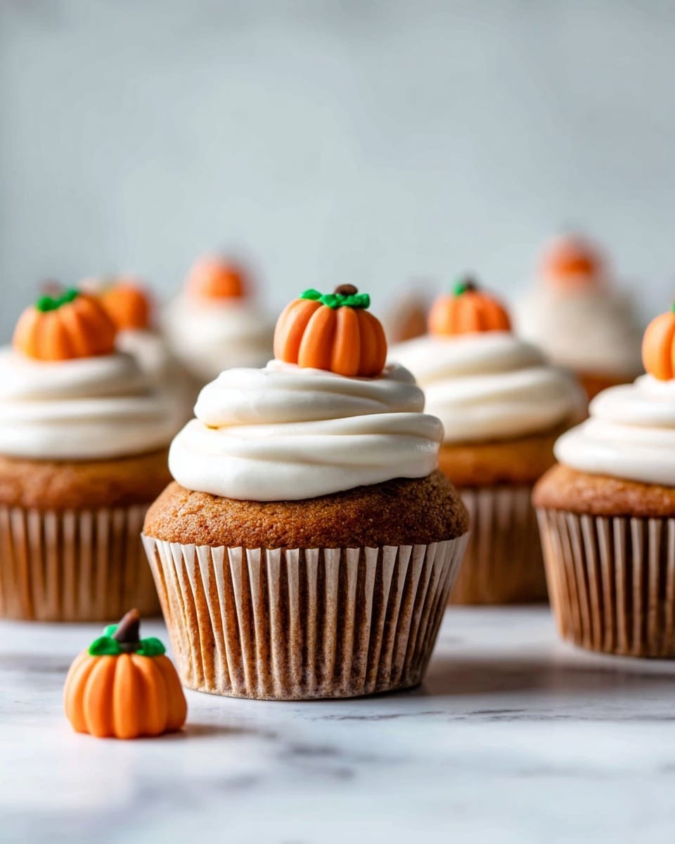 A close-up view of a small cupcake placed on a white marbled surface. The cupcake has one visible layer of soft, moist brown cake with a slightly rough texture. On top, there is a thick layer of creamy white frosting with smooth swirls and peaks. At the center of the frosting sits a small orange pumpkin decoration made of fondant or icing, detailed with green leaves and a brown stem. The cupcake is partially unwrapped from its ridged white paper liner, showing the cake's airy inside. photo taken with an iphone --ar 4:5 --v 7