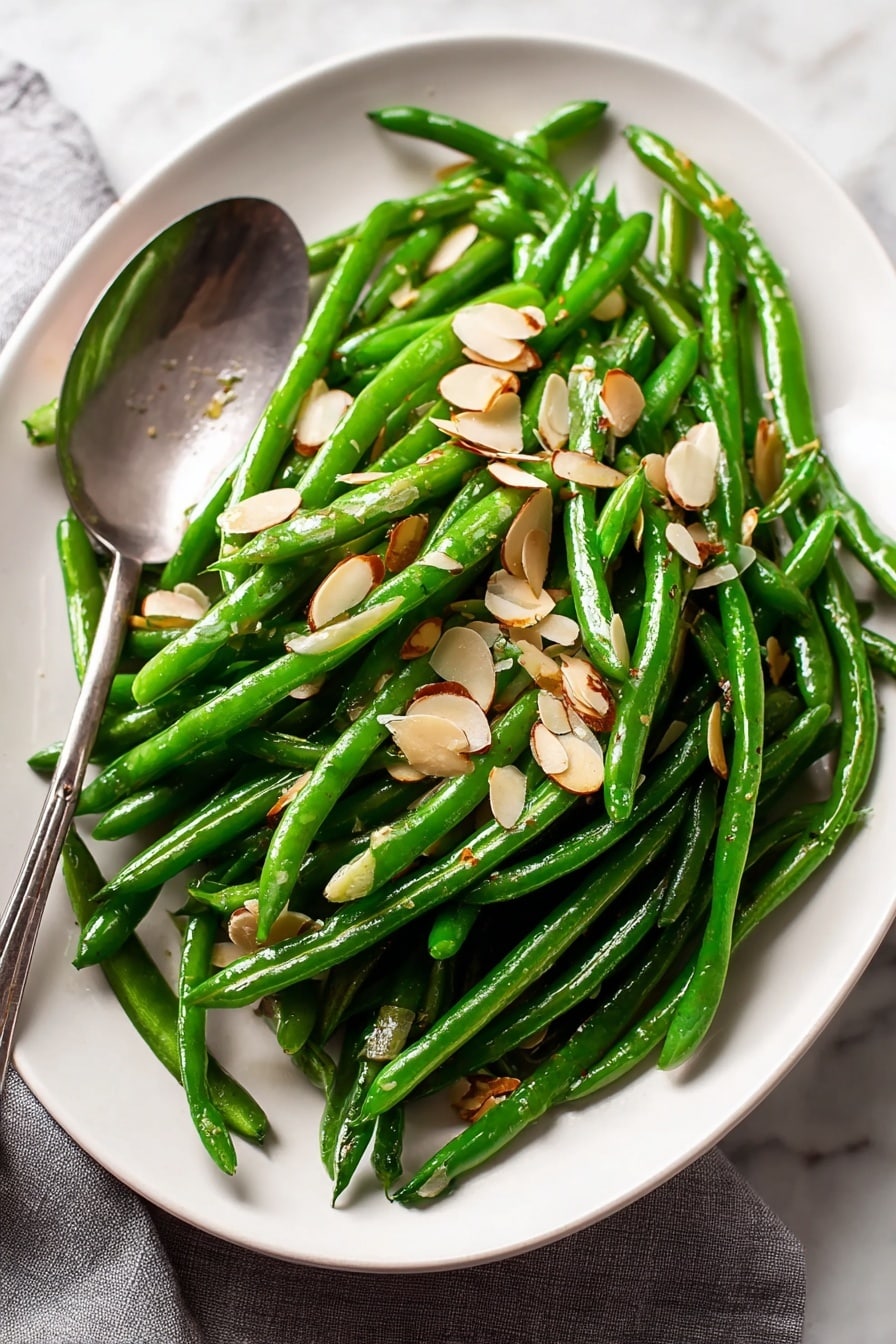 A white plate filled with bright green steamed green beans piled in a loose mound, scattered on top with thin, toasted almond slices that are light brown and slightly shiny from cooking oil. A large silver serving spoon rests on the plate under some green beans near the bottom left. The plate is placed on a white marbled surface with a grey cloth partially visible at the edge. The green beans look fresh and slightly glossy from light seasoning photo taken with an iphone --ar 2:3 --v 7 - Green Beans Almondine, Green Beans Almondine recipe, almond green beans side dish, garlic green beans with almonds, quick green bean side dish
