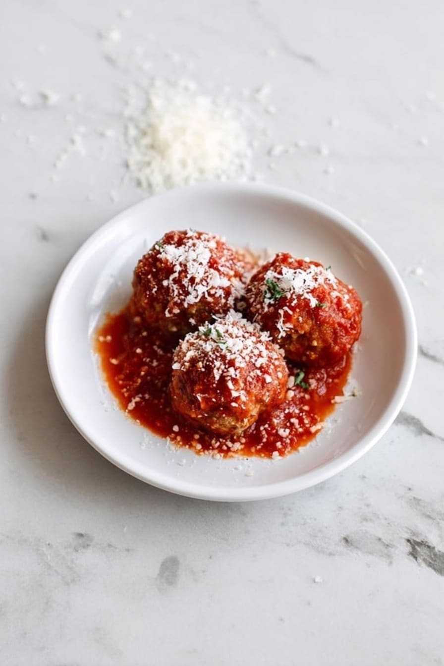 Three round meatballs covered in bright red tomato sauce are placed on a small white plate. Each meatball is topped with a white grated cheese that looks soft and crumbly. The plate sits on a white marbled surface, and to the right side, there is a wooden spoon with more white grated cheese on it. In the background, a small bunch of green herbs and a silver fork can be seen slightly blurred. The photo taken with an iphone --ar 2:3 --v 7 - Healthy Crockpot Mini Turkey Quinoa Meatballs, healthy turkey meatballs, quinoa meatballs, slow cooker turkey recipes, nutritious dinner ideas