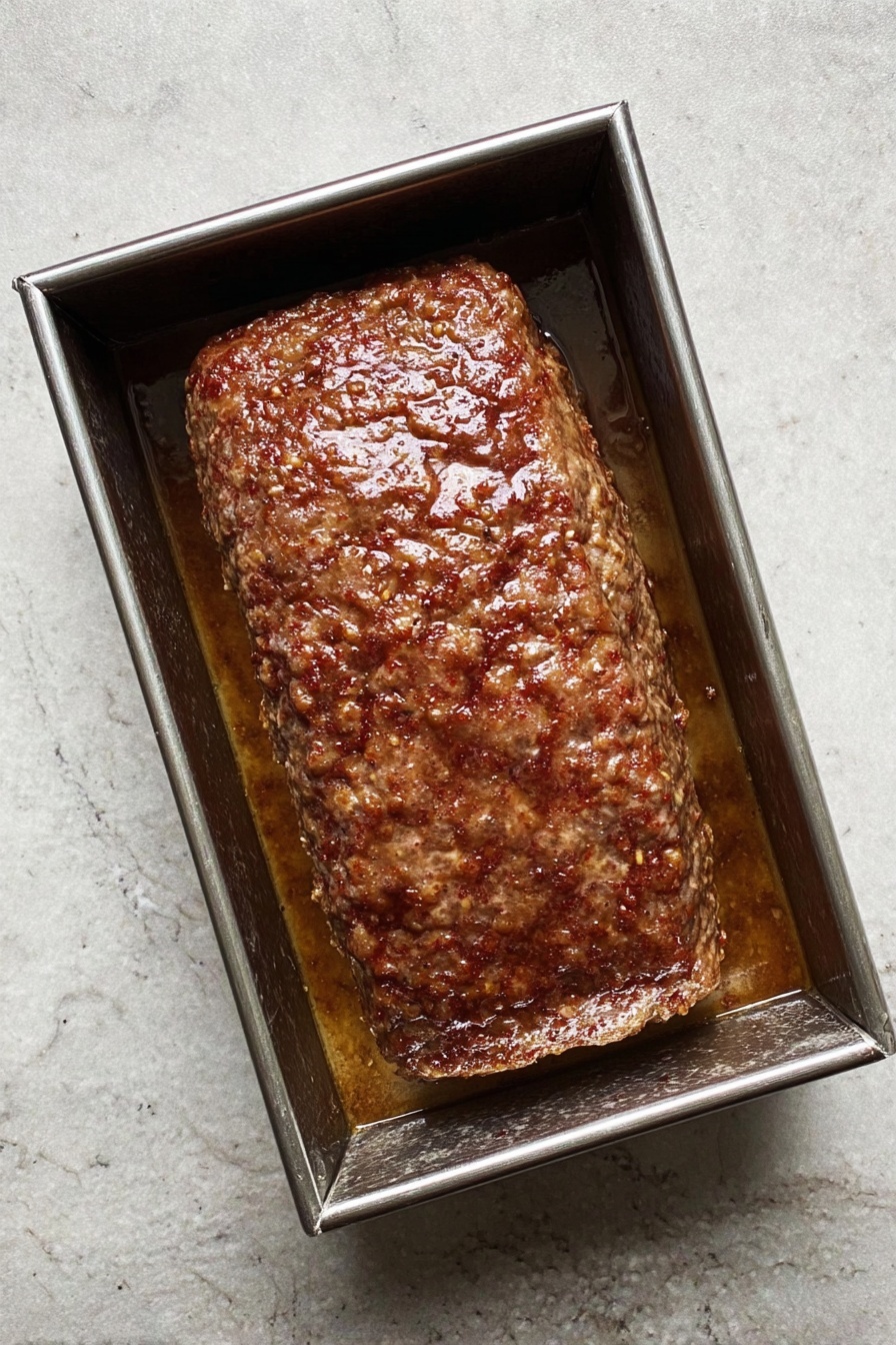A rectangular meatloaf with a browned, slightly textured crust on top sits on a wooden board. To the right of the meatloaf are five thin slices, showing a dense, even-textured inside with small dark flecks. Above the meatloaf is a large knife with a black handle placed horizontally. The background is changed to a white marbled texture. photo taken with an iphone --ar 2:3 --v 7 - Homemade Gyro Meat, gyro meat recipe, Mediterranean gyro, how to make gyro meat, homemade gyro