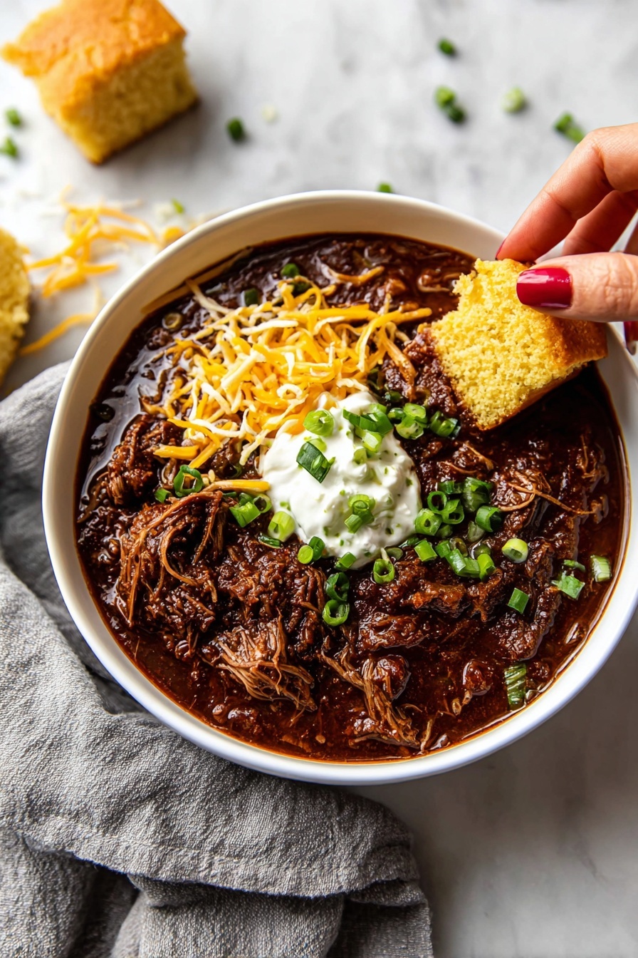 A white bowl filled with dark brown chili that has shredded meat and thick sauce at the bottom and middle layer, topped with bright orange shredded cheese and a dollop of white sour cream in the center. There are small pieces of green chopped scallions scattered on top of the chili and sour cream. A woman's hand with red-orange nail polish is holding a piece of light yellow cornbread, dipping it into the chili from the right side of the bowl. The bowl sits on a white marbled surface with a dark gray cloth partially under it and some more cornbread pieces blurred in the background. Photo taken with an iphone --ar 2:3 --v 7 - Slow Cooker Texas Beef Chili, Texas Beef Chili, hearty beef chili, easy slow cooker chili, flavorful beef chili