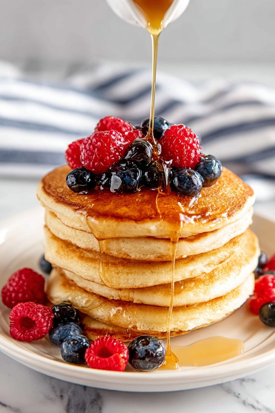 A stack of five golden brown pancakes sits in the center of a white plate, each pancake thick and fluffy with a soft texture. On top of the stack is a mix of red raspberries and dark blue blueberries, some berries spilling onto the plate around the base. A golden syrup is being poured from above, flowing down the sides of the pancakes and pooling lightly at the bottom. The plate is placed on a white marbled surface with a blurred striped cloth in the background. photo taken with an iphone --ar 2:3 --v 7 - Easy Classic Buttermilk Pancakes, fluffy pancake recipe, homemade breakfast pancakes, simple pancake batter, tender pancake recipe