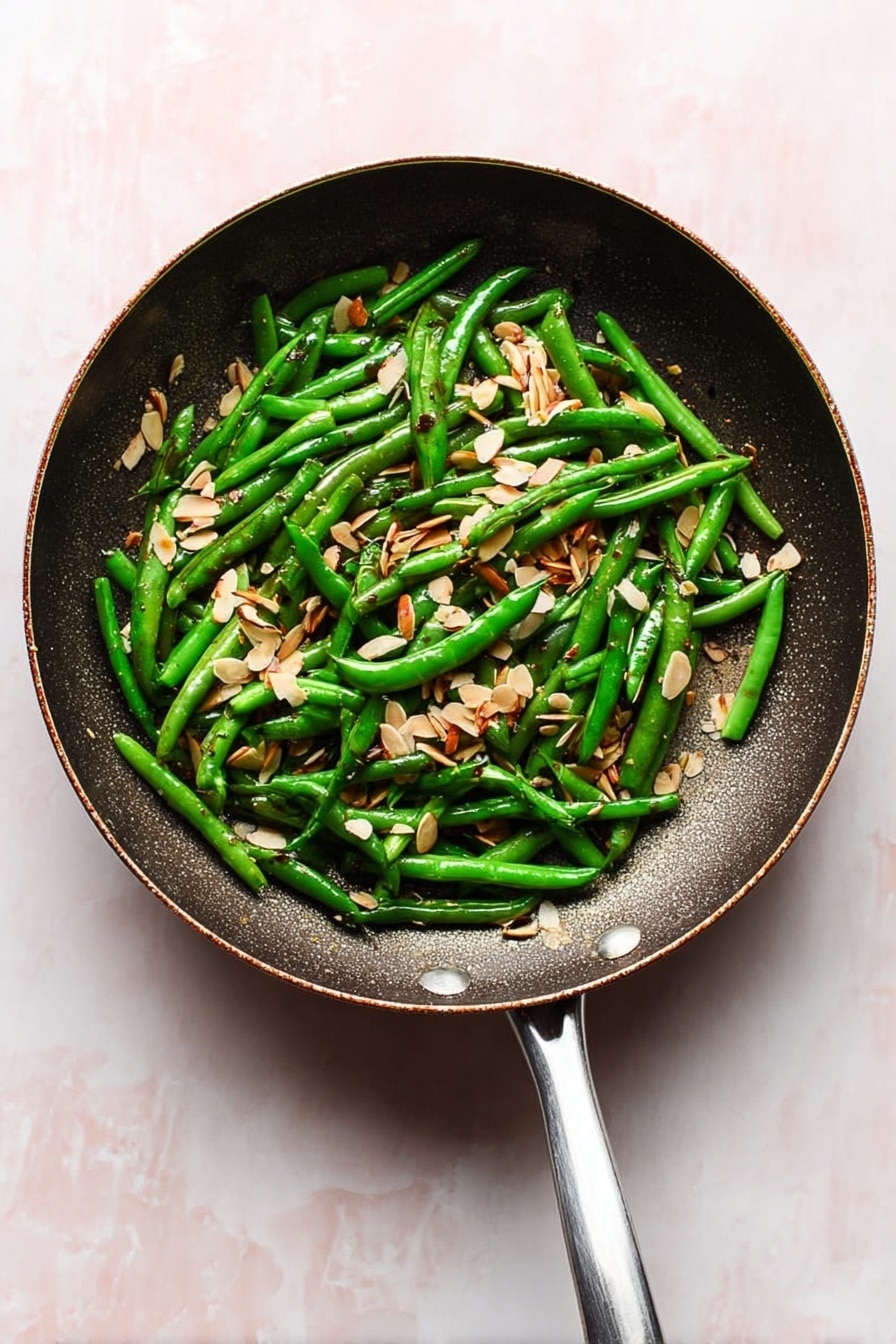 A frying pan filled with a single layer of bright green cooked green beans that are shiny, mixed evenly with scattered thin, light brown almond slices. The frying pan is dark and metallic with a central cooking surface and a silver handle extending downward. The background is a white marbled texture. photo taken with an iphone --ar 2:3 --v 7 - Green Beans Almondine, Green Beans Almondine recipe, almond green beans side dish, garlic green beans with almonds, quick green bean side dish