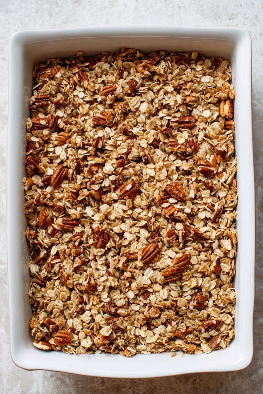 A close-up of a white rectangular baking dish filled with a single thick layer of golden brown granola, made of oats and chopped pecans, spread evenly across the dish's surface. The granola has a rough texture with toasted bits and clusters, showing a mix of light beige oat flakes and darker toasted nuts against the white marbled background. Photo taken with an iphone --ar 2:3 --v 7 - Sweet Potato Casserole with Pecans and Toasted Oat Topping, sweet potato casserole with pecans, best sweet potato casserole, holiday sweet potato dish, easy sweet potato casserole