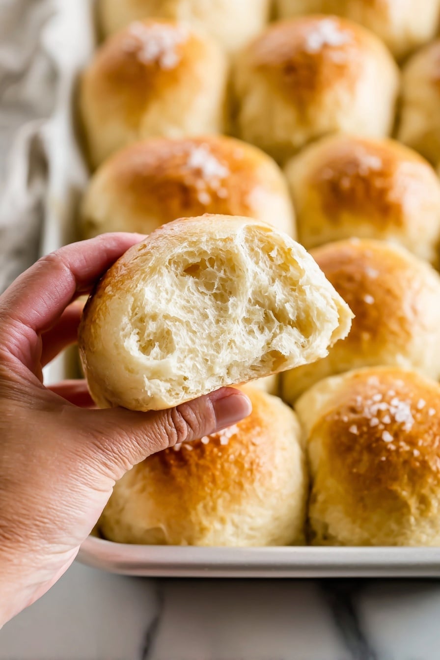 The image shows a white baking tray filled with soft, round bread rolls arranged in a close grid pattern. Each roll has a light golden-brown top with a slightly uneven surface texture and a sprinkle of coarse salt flakes on it. The bread's color is mostly pale with warm toasted spots in the center of each roll. The tray sits on a white marbled surface, and a gray and white striped cloth is partially visible to the side. photo taken with an iphone --ar 2:3 --v 7 - Homemade Honey Dinner Rolls, Fluffy Honey Dinner Rolls, Soft Dinner Rolls with Honey, Easy Honey Dinner Roll Recipe, Delicious Honey Dinner Rolls