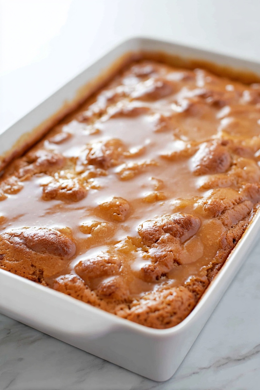 A large white rectangular baking dish holding a thick baked dessert with a golden-brown, slightly uneven cracked surface. The top is covered with a shiny, smooth glaze softly pooling into some crevices. The texture of the bake looks soft and moist with some risen, puffy areas giving it a fluffy look. The dish is placed on a white marbled surface. photo taken with an iphone --ar 2:3 --v 7 - Apple Dapple Cake with Cinnamon-Infused Glaze, apple cake with cinnamon glaze, moist apple cake recipe, cinnamon apple dessert, cozy fall cake ideas