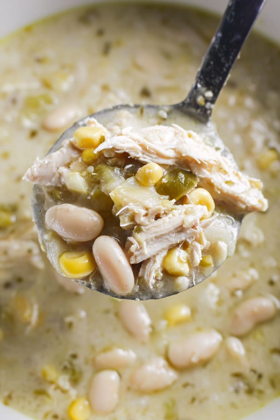 A close-up view shows a spoon filled with a thick soup containing shredded white chicken pieces, creamy white beans, light yellow corn kernels, and small green chili pieces mixed in a pale green broth. The spoon is held above the soup, which has a slightly chunky texture with visible bits of beans and chicken, all set against a white marbled texture background. photo taken with an iphone --ar 2:3 --v 7 - Green Enchilada Chicken Soup, green enchilada soup, creamy chicken soup, slow cooker chicken soup, Mexican chicken soup