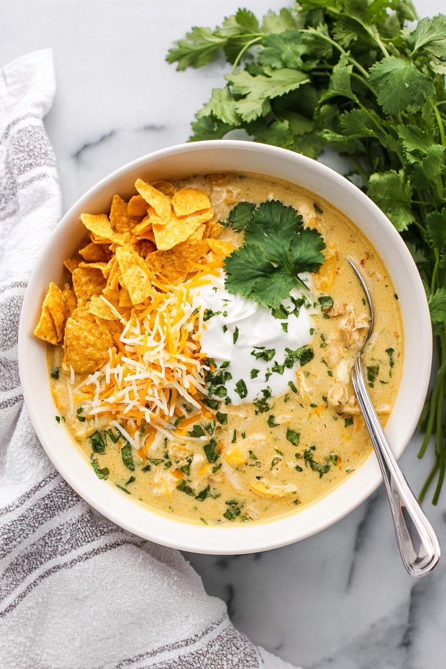 The image shows a white bowl filled with a creamy soup that has a light yellow color with green herbs mixed in. The soup is topped with three main layers: on the left side, there are broken yellow tortilla chips, on the middle bottom, there is a heap of shredded cheese in white and orange colors, and on the right middle, a dollop of white sour cream sprinkled with green herbs. A large cilantro leaf is placed on top of the chips and cheese. A silver spoon is resting inside the bowl on the right side. The bowl is placed on a white marbled surface with fresh cilantro visible in the background, and a white towel with grey stripes is partially visible on the left side. Photo taken with an iphone --ar 2:3 --v 7 - Green Enchilada Chicken Soup, green enchilada soup, creamy chicken soup, slow cooker chicken soup, Mexican chicken soup
