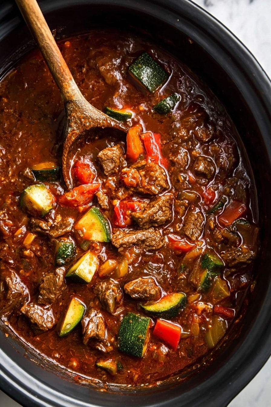 This image shows a thick stew in a black cooking pot with a wooden spoon resting inside. The stew has multiple layers of rich, dark brown sauce mixed with chunks of tender beef, pieces of green zucchini, small red bell pepper slices, and bits of onion scattered throughout. The stew looks hearty with visible oil glistening on the surface and a textured mix of soft and chunky ingredients. The pot sits on a white marbled surface. photo taken with an iphone --ar 2:3 --v 7 - Crock-Pot Paleo Beef Chili, healthy beef chili, slow cooker paleo chili, low carb beef chili, hearty paleo dinner