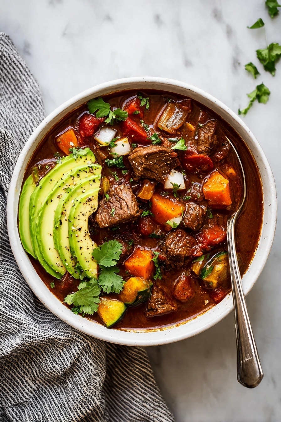 A white bowl filled with a rich dark brown stew that has chunky pieces of beef and bright colored vegetables like yellow squash, red bell peppers, and green zucchini. On the left side of the bowl, there are three thin slices of light green avocado sprinkled with black pepper. Small white onion pieces are scattered over the top along with fresh green cilantro leaves. A silver spoon rests inside the bowl on the left edge. The bowl is placed on a white marbled surface. photo taken with an iphone --ar 2:3 --v 7 - Crock-Pot Paleo Beef Chili, healthy beef chili, slow cooker paleo chili, low carb beef chili, hearty paleo dinner