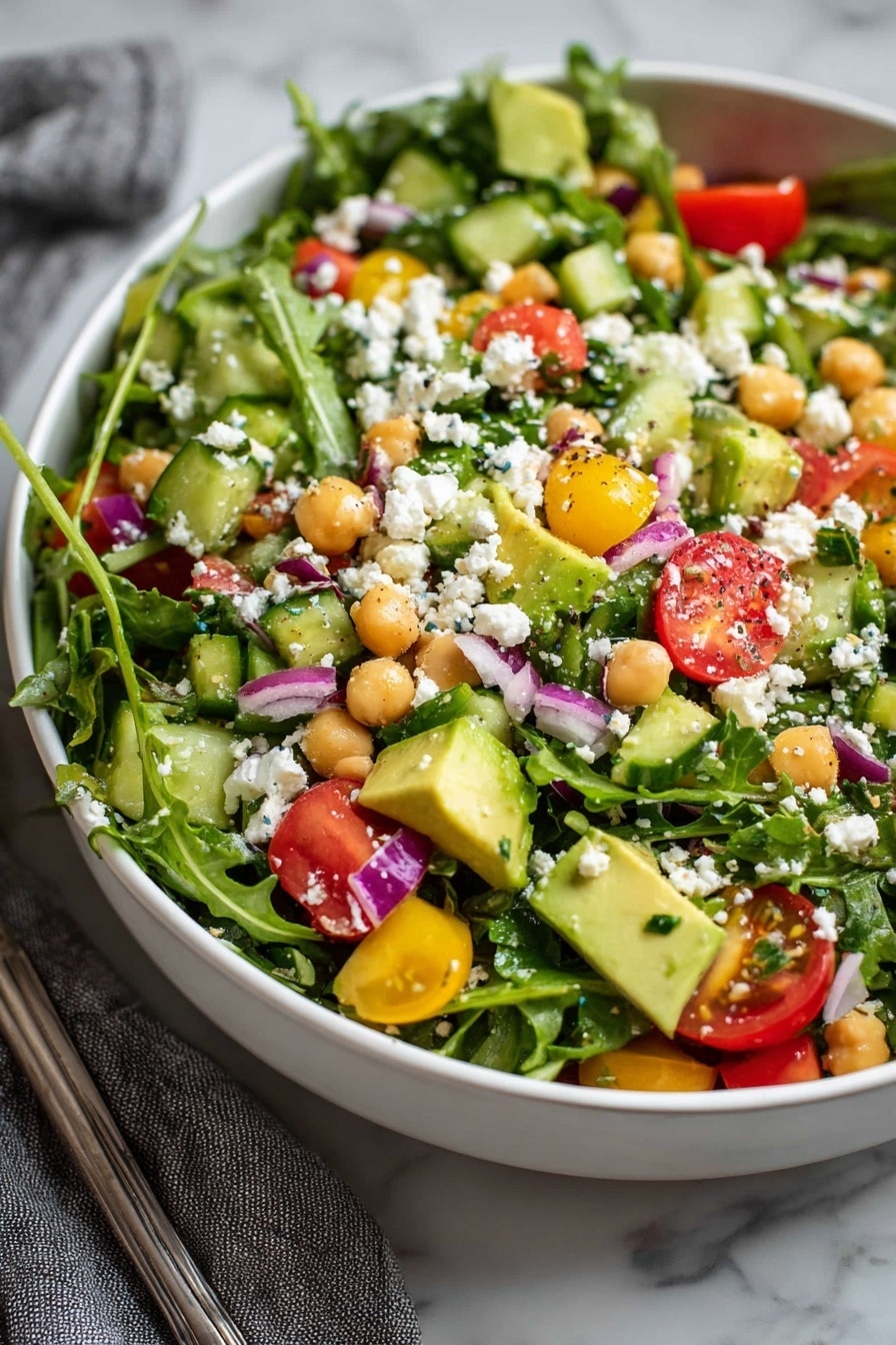 A close-up view of a fresh salad in a white bowl on a white marbled surface, showing mixed green arugula leaves as the base layer with a few chickpeas scattered through. On top, there is a mix of bright red halved cherry tomatoes and light green cucumber slices with a crunchy texture. Small chunks of creamy white cheese are sprinkled all over, contrasting with the purple bits of onion. Black-tipped gold and black salad tongs rest inside the bowl, partially covered by the salad leaves. Photo taken with an iphone --ar 2:3 --v 7 - Chickpea Avocado Salad, healthy chickpea salad, easy avocado salad, vegan chickpea recipes, nutritious lunch salad