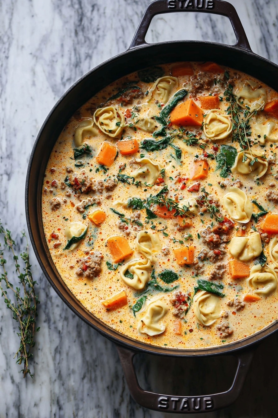 A pan filled with creamy soup that has three main layers: a light orange creamy broth with small oil spots on top, floating bright orange chunks of squash, and pale yellow tortellini pasta. The soup also has bits of cooked ground meat spread evenly and green spinach leaves and fresh thyme sprigs scattered on the surface, giving a mix of colors and textures in each spoonful. The pan is black with a handle that has the word “STAUB” on it. The background is a white marbled texture. photo taken with an iphone --ar 2:3 --v 7 - Creamy Sausage Tortellini Soup, Sausage Tortellini Soup, comforting creamy soup recipes, hearty sausage soup, easy weeknight soup ideas
