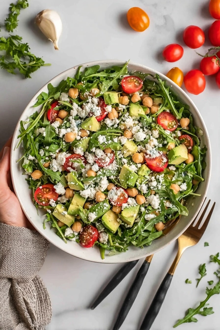 A white bowl filled with a fresh, colorful salad sits on a white marbled surface, layered with dark green arugula leaves as the base. Mixed throughout are small yellow avocado chunks, light beige chickpeas, bright red cherry tomato halves, and light green cucumber pieces. Crumbled white feta cheese is sprinkled evenly on top, along with small pieces of purple onion, adding texture and contrast. Two black and wooden salad forks rest inside the bowl to the right, and a white and gray striped cloth lies next to it on the surface. Photo taken with an iphone --ar 2:3 --v 7 - Chickpea Avocado Salad, healthy chickpea salad, easy avocado salad, vegan chickpea recipes, nutritious lunch salad