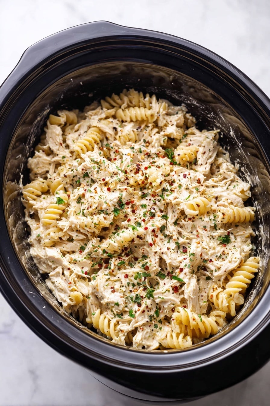 A close-up of a wooden spoon lifting a creamy pasta dish from a black pot. The pasta is rotini, coated in a thick white sauce with visible shredded pieces of chicken mixed in. The sauce is speckled with green herbs and red chili flakes, giving a hint of color contrast. The black pot holds more of the creamy pasta mixture, with a soft, slightly grainy texture from the sauce and spices. The background shows a white marbled surface. Photo taken with an iphone --ar 2:3 --v 7 - Crockpot Garlic Parmesan Chicken Pasta, slow cooker chicken pasta, creamy garlic chicken pasta, easy crockpot chicken recipe, comforting chicken pasta dish