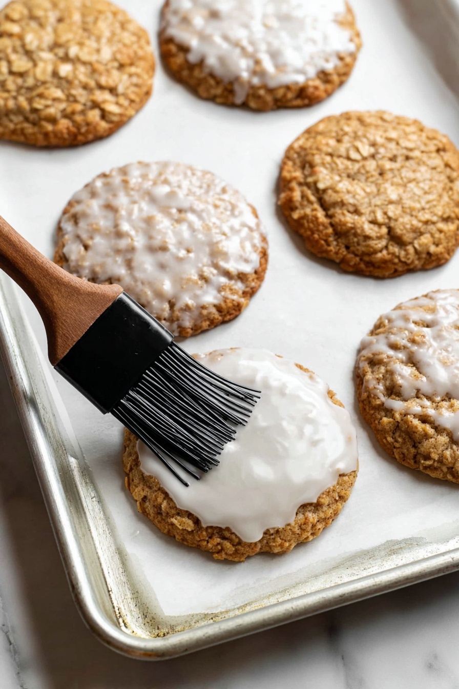 A close-up of a silver baking tray lined with white parchment paper holding five large oatmeal cookies arranged in a loose pattern, with two cookies topped with a glistening white icing glaze and the others plain showing rough oat textures; a black pastry brush spreading the shiny icing over the central cookie; all sitting on a white marbled surface photo taken with an iphone --ar 2:3 --v 7 - Gingerbread Oatmeal Cookies, gingerbread cookies, oatmeal cookies, holiday cookies, spiced cookie recipe