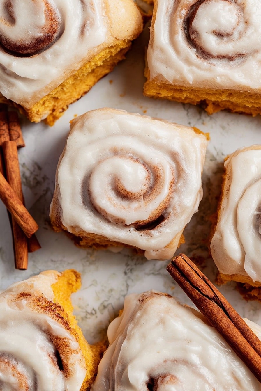 The image shows two cinnamon rolls lined up vertically on parchment paper. Each roll has multiple layers of golden brown dough spiraled around a darker cinnamon filling. The top of the rolls is covered with a thick, creamy white icing that is slightly melting and spreading down the sides. The icing has a smooth texture with hints of cinnamon sprinkled on top. In the background, there are two cinnamon sticks crossed on a white marbled surface. photo taken with an iphone --ar 2:3 --v 7 - Pumpkin Cinnamon Rolls with Cream Cheese Icing, fall pumpkin cinnamon rolls, easy pumpkin cinnamon rolls, fluffy pumpkin cinnamon rolls, pumpkin spice cinnamon rolls
