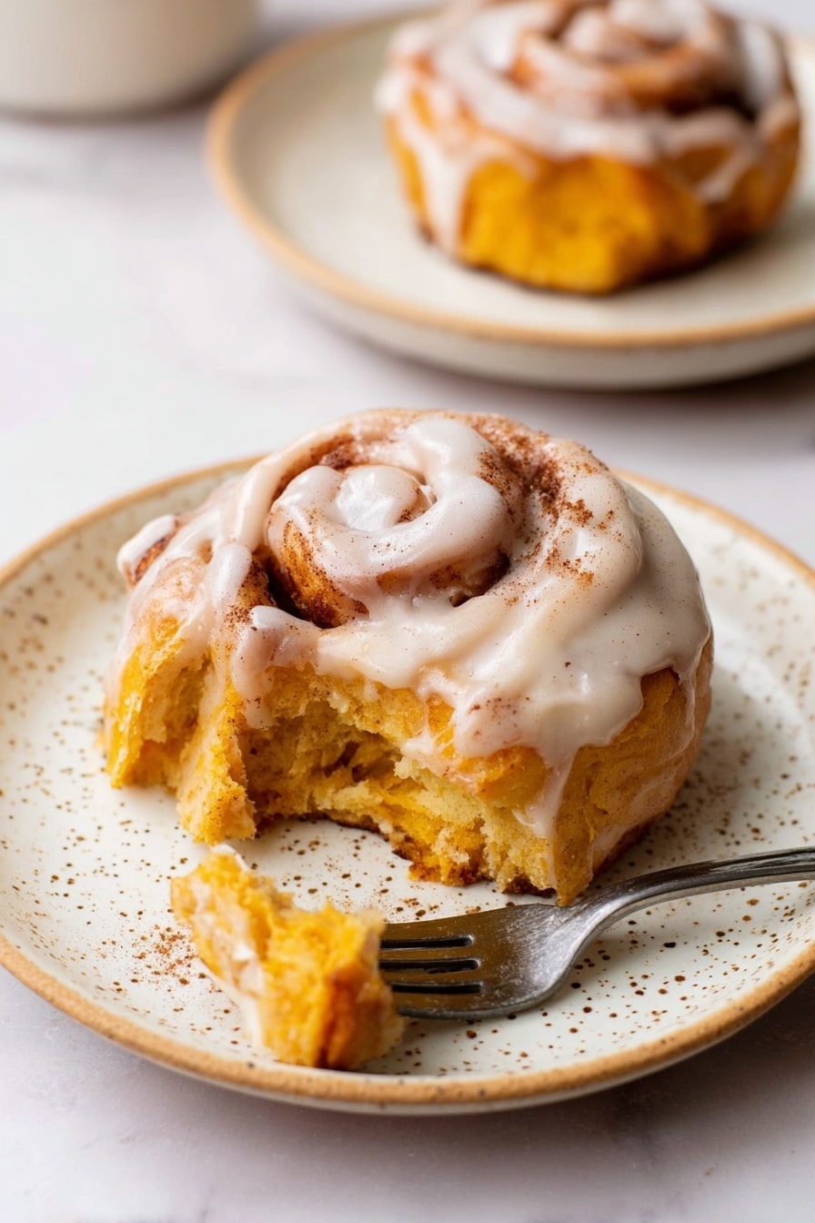 A single cinnamon roll with two visible layers of soft, golden orange dough swirled around each other sits on a white speckled plate, topped with a thick, creamy white glaze that covers the top and drips slightly down the sides, showing a dusting of cinnamon on the glaze. A small piece is torn from the roll and resting on a silver fork at the plate's edge, revealing the fluffy texture inside. In the background, another similar roll on a white speckled plate stands out on a white marbled surface. Photo taken with an iphone --ar 2:3 --v 7 - Pumpkin Cinnamon Rolls with Cream Cheese Icing, fall pumpkin cinnamon rolls, easy pumpkin cinnamon rolls, fluffy pumpkin cinnamon rolls, pumpkin spice cinnamon rolls