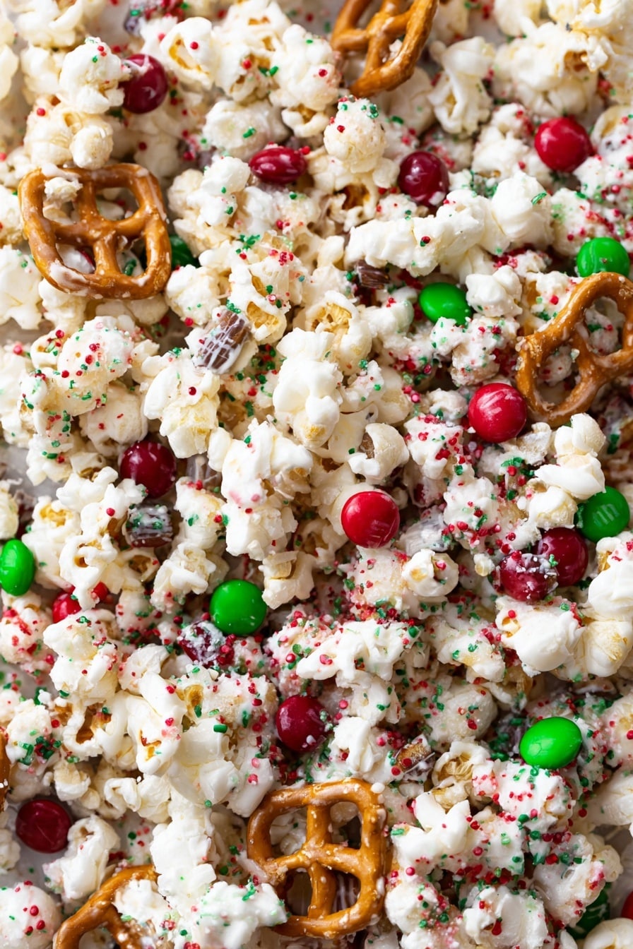 The image shows a mix of white popcorn pieces coated with a light white drizzle that looks like white chocolate, scattered with small red, green, and white round and square sprinkles. Among the popcorn are golden brown pretzels with a shiny surface, and round red and green candy-coated chocolates, creating a colorful and festive look. All the ingredients are close together in a heap that fills the frame. The background is a white marbled texture. photo taken with an iphone --ar 2:3 --v 7 - White Chocolate Christmas Popcorn, festive holiday popcorn, sweet and salty holiday snacks, easy holiday popcorn ideas, Christmas party treats