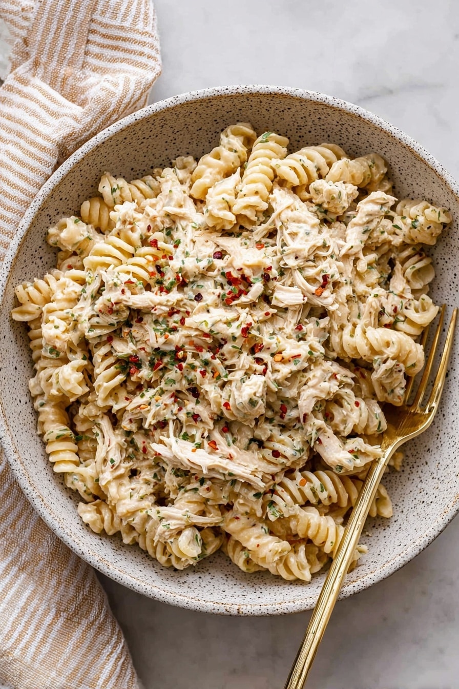 A large white speckled bowl filled with creamy pasta made of spiral-shaped noodles mixed with shredded chicken. The creamy sauce covers the pasta and chicken, showing bits of herbs and small red pepper flakes scattered on top. A silver fork with a golden handle rests on the right side of the bowl, partly submerged in the pasta. The bowl sits on a white marbled surface with a beige and white striped cloth napkin placed beside it. Photo taken with an iphone --ar 2:3 --v 7 - Crockpot Garlic Parmesan Chicken Pasta, slow cooker chicken pasta, creamy garlic chicken pasta, easy crockpot chicken recipe, comforting chicken pasta dish