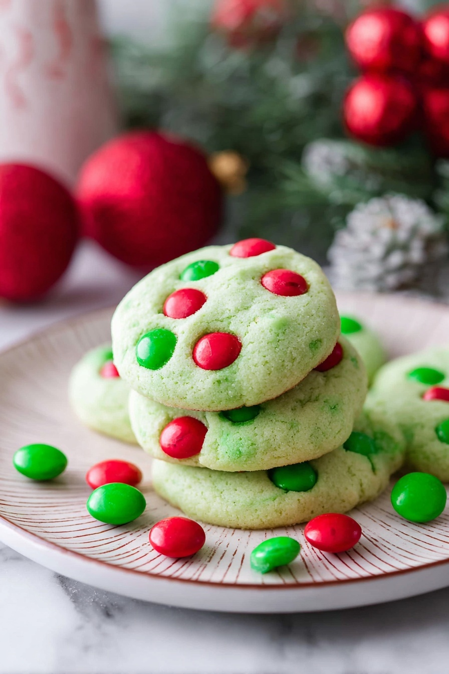 Eight round green cookies with red and green candy pieces on top are placed on a metal cooling rack in the center of a white marbled surface. One cookie is broken into two halves at the bottom left. Red and green candy pieces are scattered around the rack. A glass jar filled with more red and green candies is at the right edge. Some green leaves are partially visible in the bottom left corner. The cookies have a soft, slightly cracked texture with candies sunken into the dough. Photo taken with an iphone --ar 2:3 --v 7 - Grinch Cookies with M&Ms, festive Christmas cookies, holiday baking ideas, colorful holiday cookies, easy Grinch cookie recipe