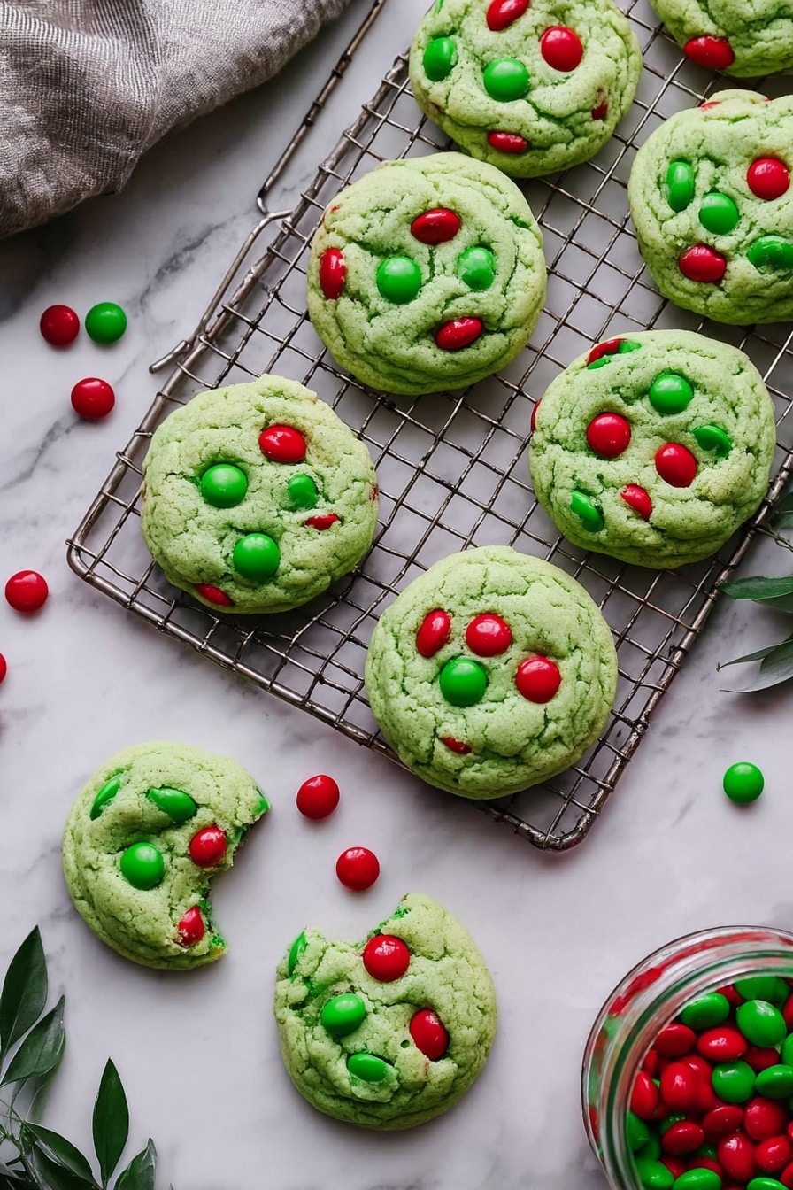 The image shows a stack of four soft, thick green cookies with a smooth texture, each studded with bright red and green candy-coated chocolates pressed into the top surfaces. The top cookie is broken in half, displaying a moist, dense green inside. The cookies sit on a dark metal cooling rack placed over a white marbled surface. Around the stack, more green cookies with red candies and a few loose red and green candies scatter on the surface. The background is softly blurred with hints of festive colors and greenery. Photo taken with an iphone --ar 2:3 --v 7 - Grinch Cookies with M&Ms, festive Christmas cookies, holiday baking ideas, colorful holiday cookies, easy Grinch cookie recipe