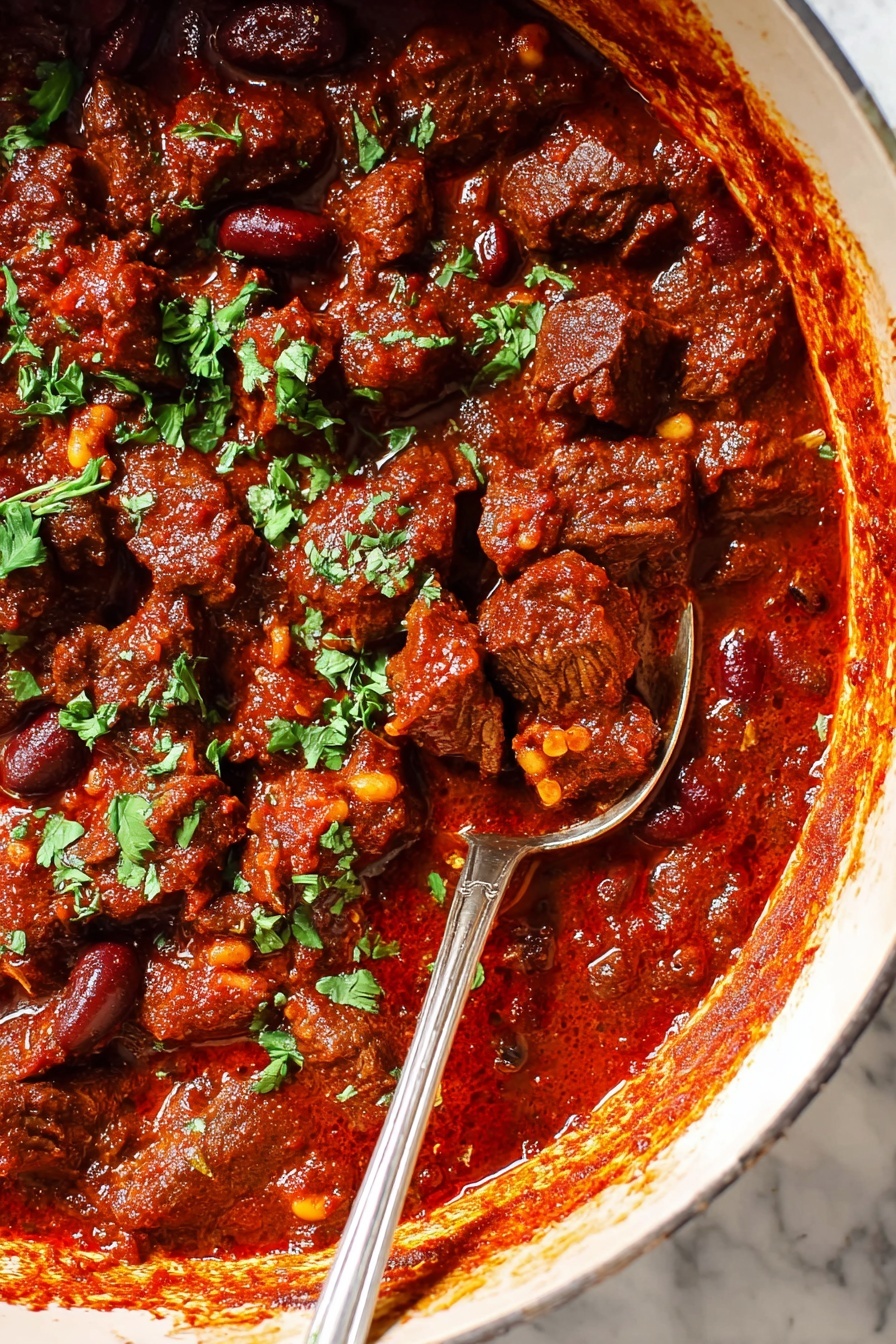The image shows a close-up of a thick red stew filled with chunks of dark brown meat and small kidney beans, covered in a rich, reddish sauce with a slightly glossy texture. There are small bits of fresh green herbs sprinkled evenly on top, adding a pop of color. A silver spoon with a long handle is partially submerged on the right side, lifting some meat and sauce. The stew is inside a large white pot with a hint of its rim visible, sitting on a white marbled surface. Photo taken with an iphone --ar 2:3 --v 7 - Hearty Beef Chili Con Carne, Beef Chili Recipe, Easy Chili Con Carne, Comforting Beef Chili, Slow-Cooked Beef Chili