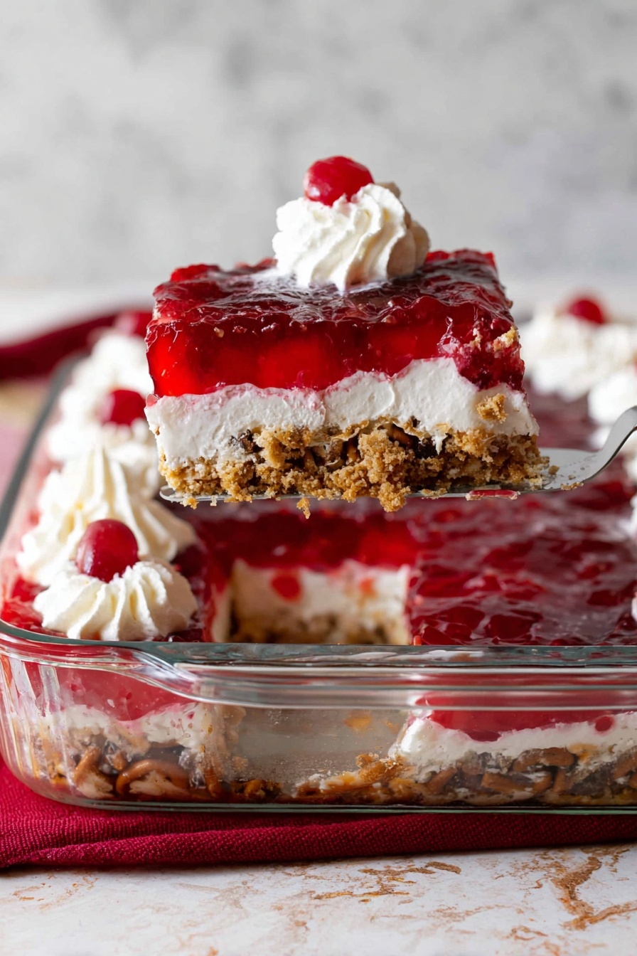 This image shows a clear glass baking dish with a dessert made of four layers. The bottom layer is a light brown crumbly crust with visible pieces of nuts or pretzels. Above it is a thick white creamy layer. The third layer is a bright red jelly with pieces of cherries inside. The top has several small white whipped cream swirls, with one slice being lifted by a spatula. The slice has a dollop of whipped cream and a single cherry on top. The background and surface show a white marbled texture, with a red cloth partially visible in the front. Photo taken with an iphone --ar 2:3 --v 7 - Cranberry Pretzel Salad, easy cranberry pretzel salad, holiday pretzel dessert, layered cranberry salad, salty sweet pretzel salad