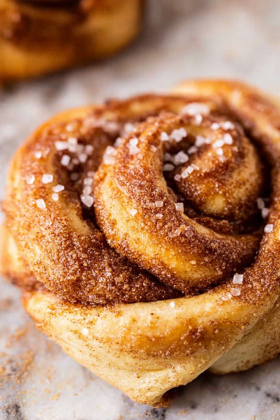 The image shows a close-up of a cinnamon roll with two visible layers: the outer layer is light golden brown dough twisted in a spiral, coated with a layer of cinnamon and sugar, while the inner layer reveals a darker, rich cinnamon filling. Large sugar crystals are sprinkled over the top, giving a crunchy texture alongside the soft dough. The background is a white marbled surface with a slight blur that highlights the cinnamon roll's texture and detail. Photo taken with an iphone --ar 2:3 --v 7 - Pumpkin Spice Danish Twists, pumpkin spice pastry, fall Danish pastries, easy pumpkin spice dessert, holiday baking recipes