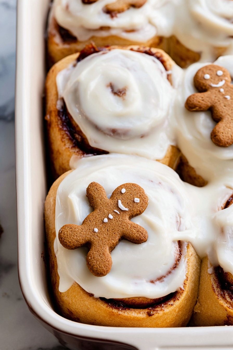 The image shows a close-up of a white baking dish filled with three cinnamon rolls covered in smooth, creamy white icing with tiny specks of cinnamon visible in the frosting. The cinnamon rolls have a golden-brown color with dark brown swirls of cinnamon in the middle. Two small gingerbread cookies, shaped like little people, are placed on top of the rolls, adding a touch of ginger spice color with their warm brown shade. The baking dish sits on a white marbled surface. photo taken with an iphone --ar 2:3 --v 7 - Gingerbread Cinnamon Rolls with Brown Butter Icing, festive gingerbread cinnamon rolls, holiday cinnamon roll recipe, spiced gingerbread rolls with cream cheese frosting, homemade gingerbread cinnamon buns