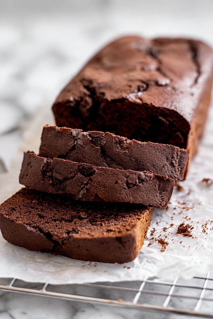 A rectangular chocolate loaf cake with a cracked top sits on white parchment paper over a silver metal cooling rack on a white marbled surface. Two thick slices lie in front of the loaf, showing a dark, moist interior with small chunks of nuts or chocolate. The cake’s surface is slightly rough and textured, with a few crumbs scattered around the sliced pieces photo taken with an iphone --ar 2:3 --v 7 - Double Chocolate Loaf Cake, chocolate loaf cake, easy chocolate cake recipe, moist chocolate cake, quick chocolate dessert