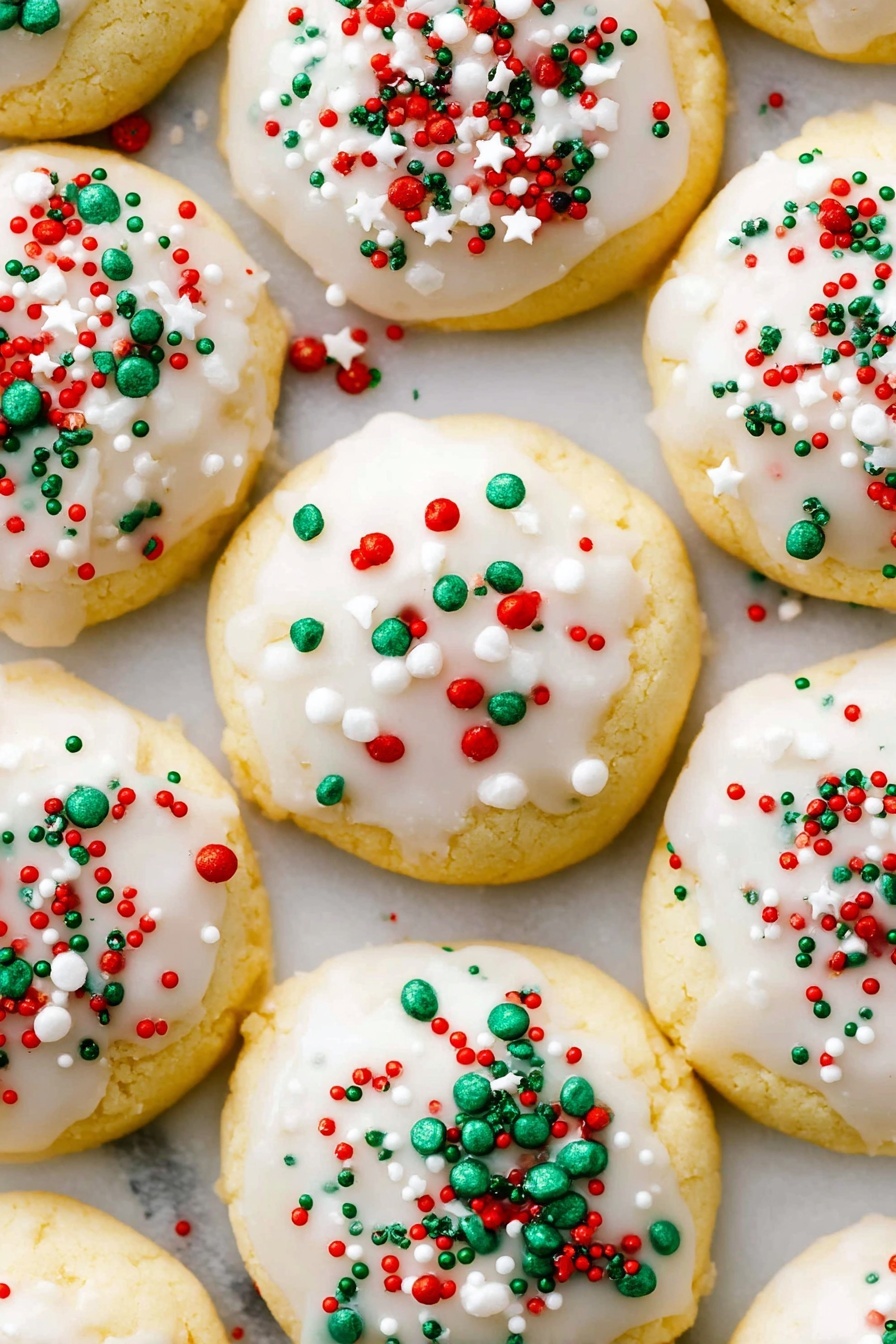 A white plate holds a pile of small round cookies that have a light golden-yellow base layer with a smooth, white icing layer on top. Each cookie is sprinkled with tiny round and rod-shaped sprinkles in red, green, and white colors, scattered unevenly on the icing. The cookies are stacked close together, with some overlapping, set against a white marbled surface background. Photo taken with an iphone --ar 2:3 --v 7 - Italian Ricotta Christmas Cookies, holiday Italian cookies, soft Christmas cookies, almond glaze cookies, festive holiday cookies
